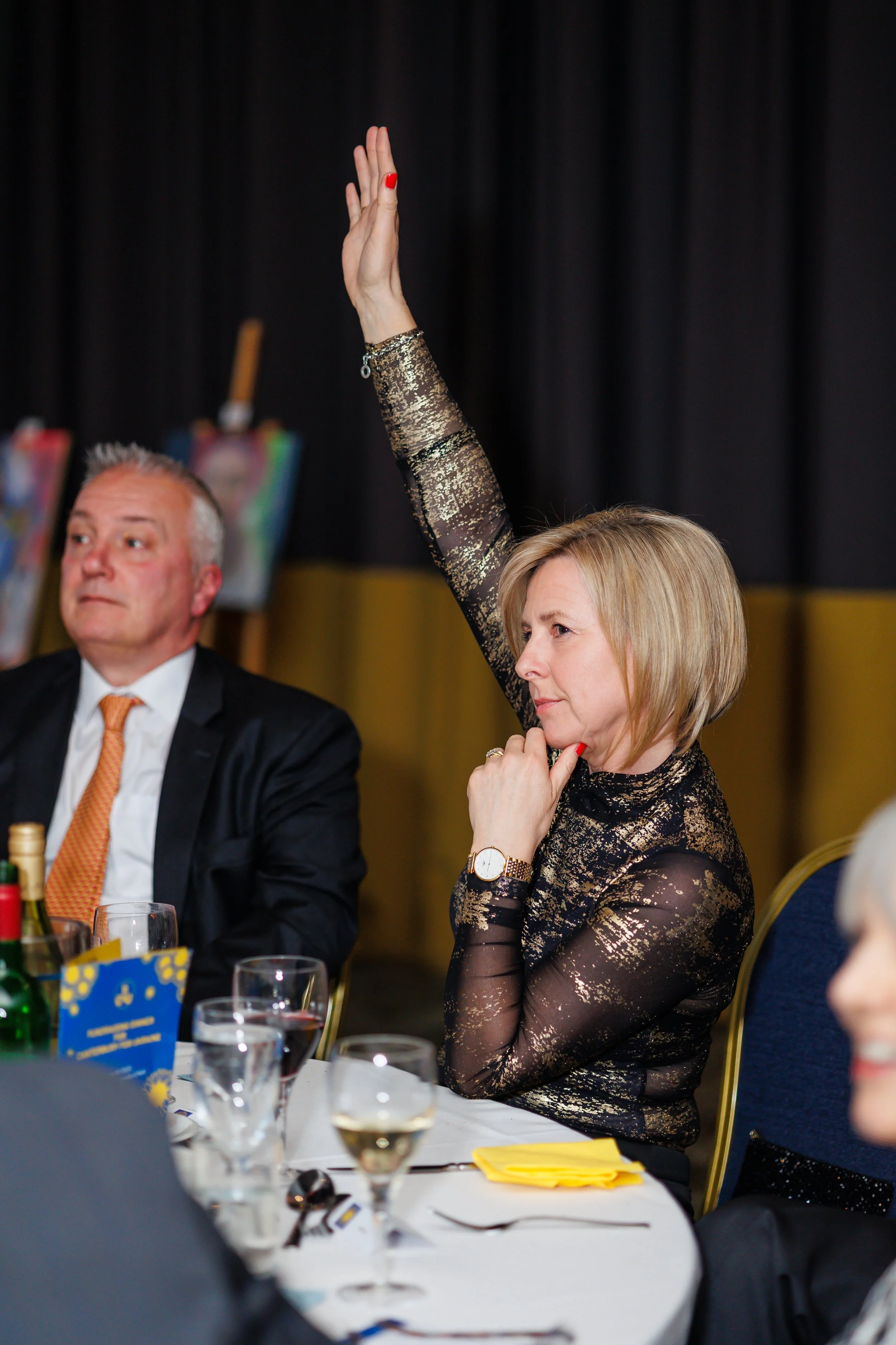 A woman with blonde hair and a gold and black patterned dress raising her hand during an auction at a Ukrainian fundraiser event at University of Kent, seated at a table with wine glasses and yellow napkins, with a man in a suit sitting beside her.