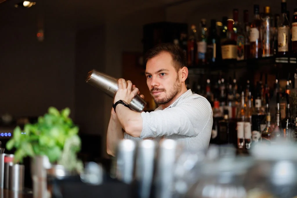 A bartender shaking a cocktail shaker behind the bar in a dimly lit bar area, with bottles of alcohol on shelves in the background at the Rocksalt in Folkestone.