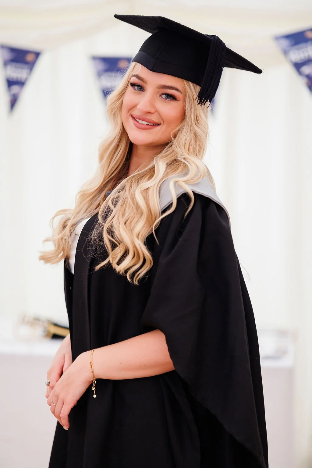 Young woman with long wavy blonde hair in graduation cap and gown smiling at camera during a University of Kent in Canterbury graduation celebration.