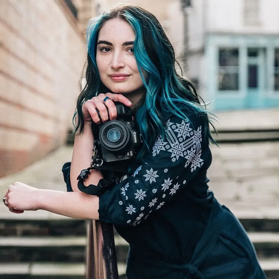 Young woman with blue and black hair holding a camera, wearing a dark sweater with white snowflake patterns, standing outdoors in front of a brick wall and stairs.