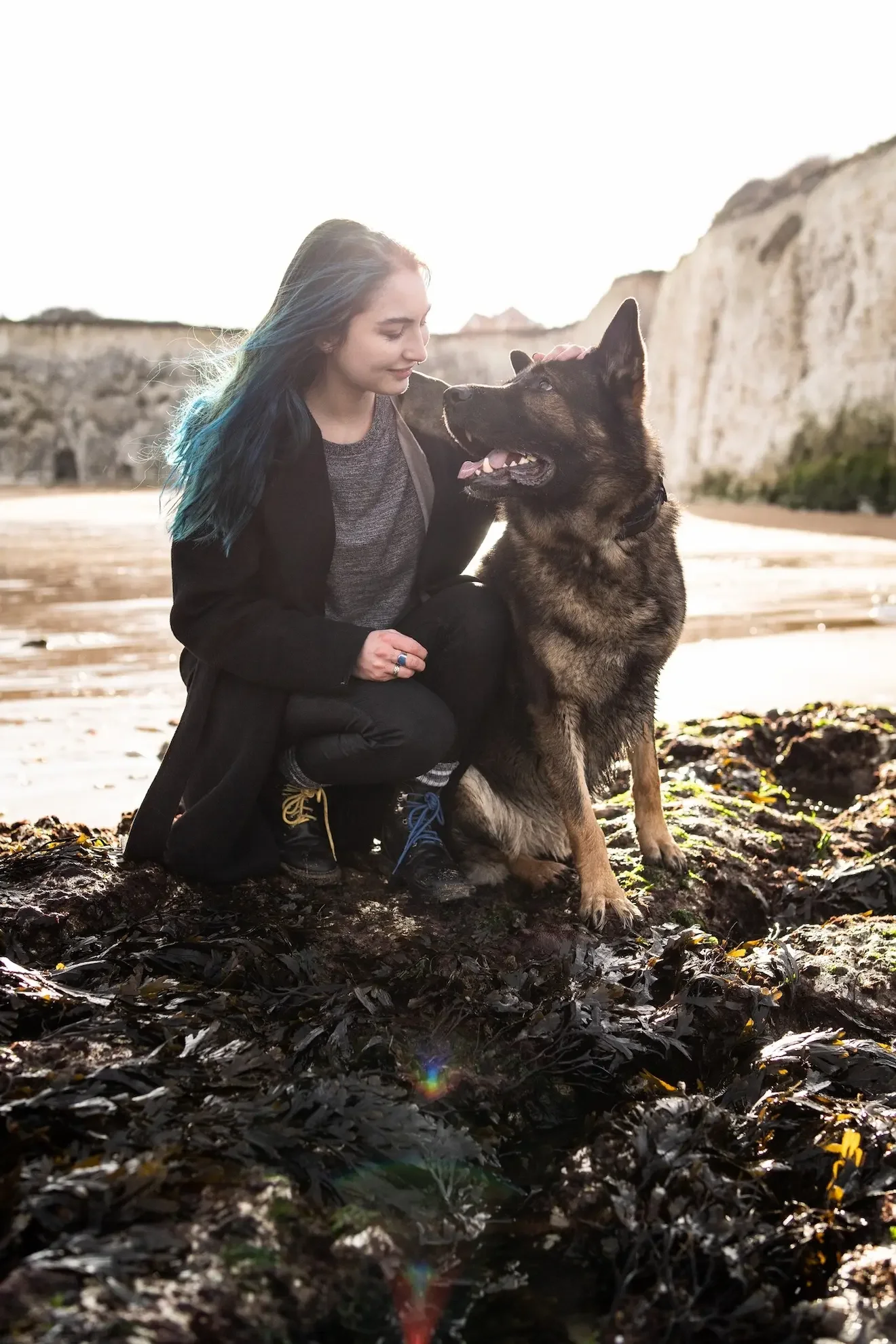 A woman crouching on a rocky beach with seaweed, looking at a large German Shepherd dog, with white cliffs in the background during sunset