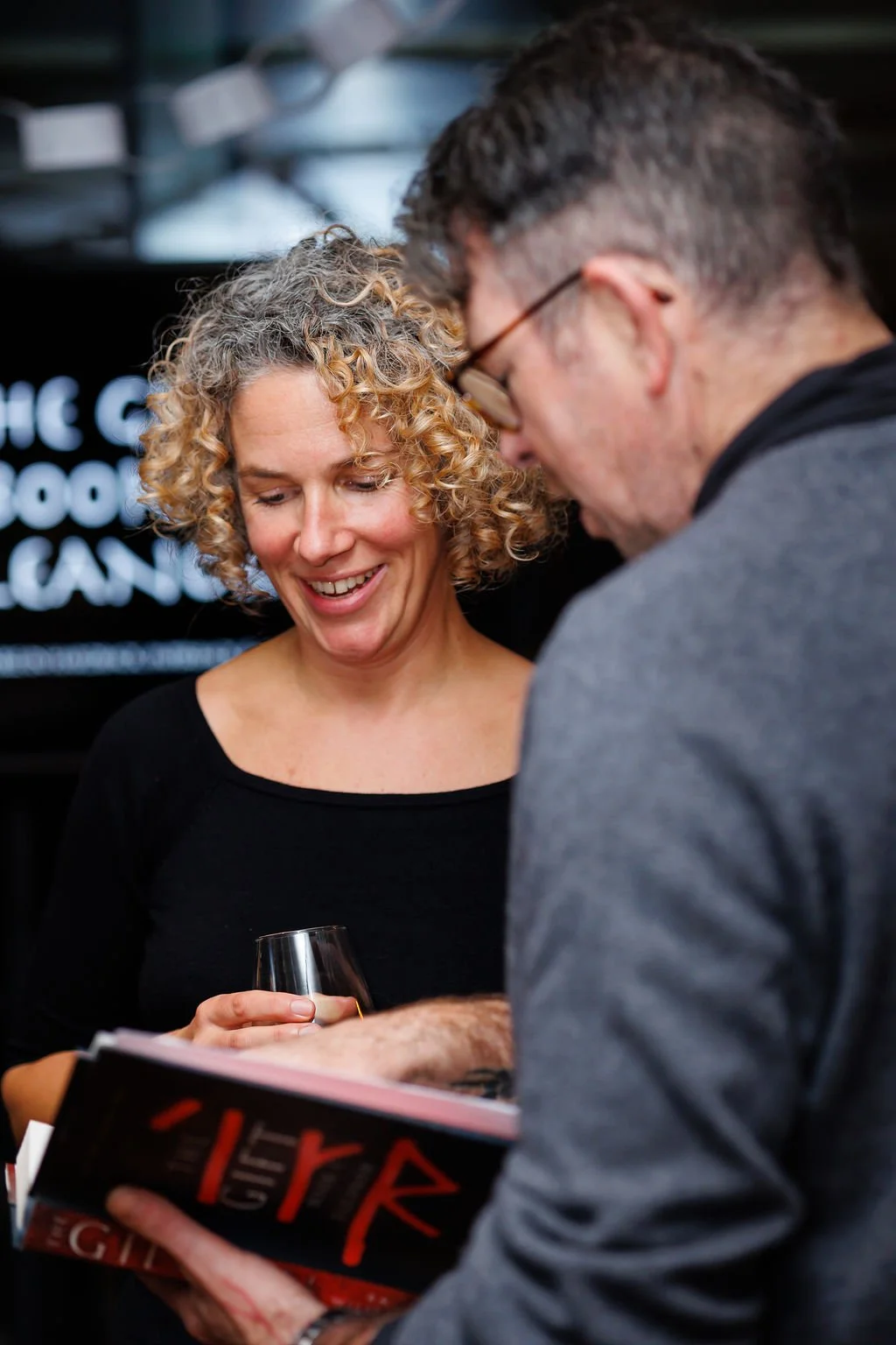 A woman with curly blonde hair smiling and holding a wine glass, looking at a man with glasses who is signing a book, at a book launch event at Rocksalt in Folkestone during Book Festival.