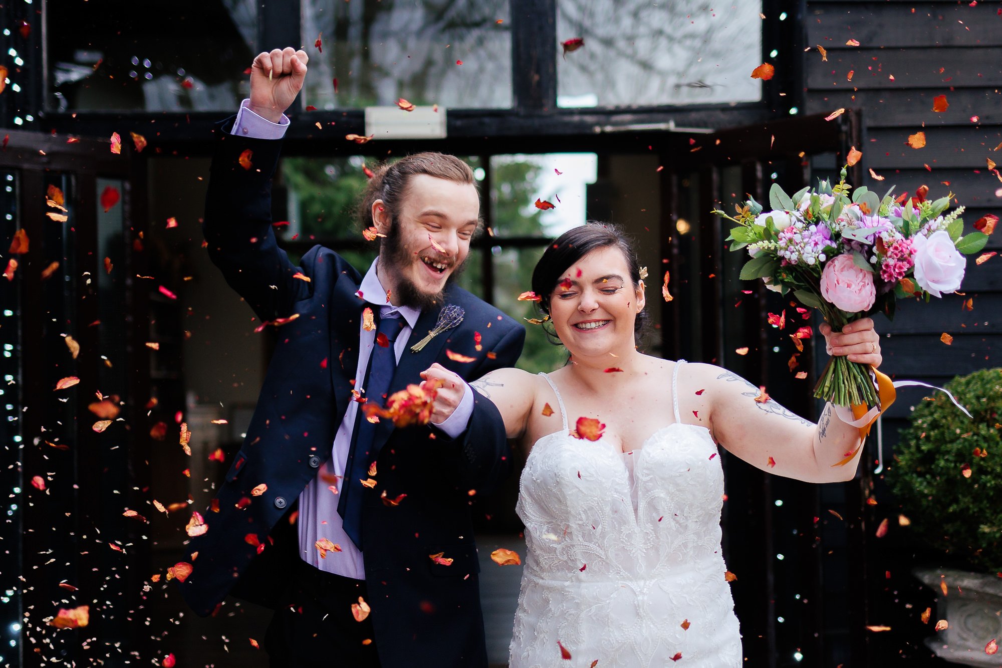 A newly married couple celebrating their wedding outdoors, surrounded by falling flower petals. The groom, with a beard and long hair tied back, is wearing a dark suit with a white shirt and tie. The bride, with short dark hair, is in a white lace wedding dress holding a bouquet of pink and purple flowers. Both are smiling and joyful.