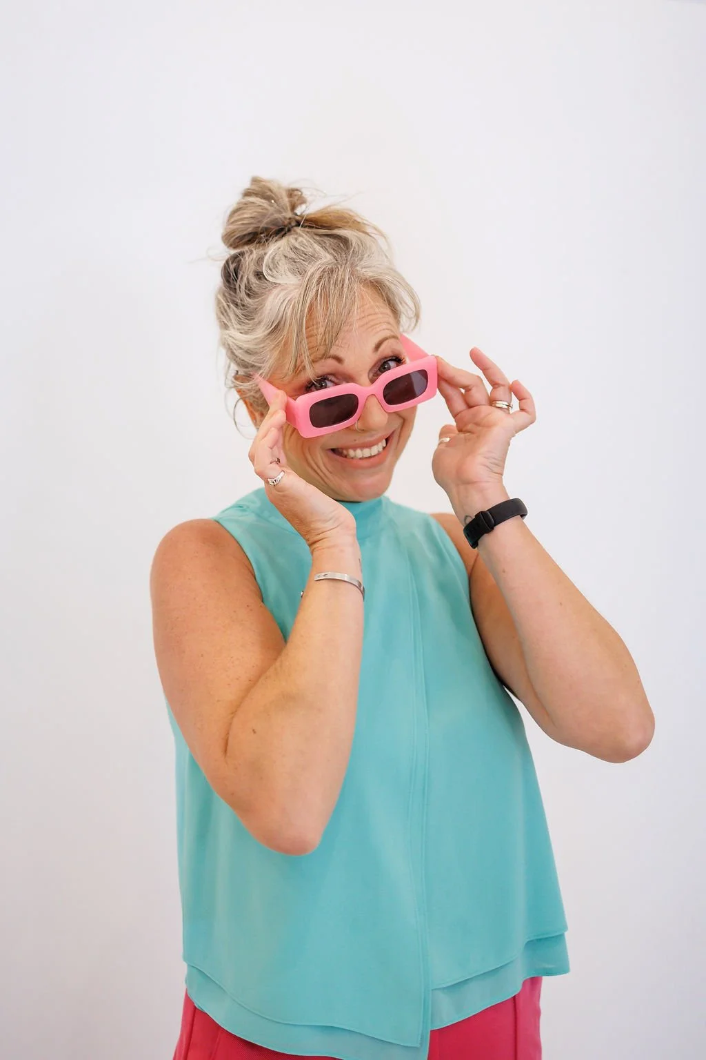 A woman with gray hair styled in a messy bun, wearing a light blue sleeveless top, pink sunglasses, and a black watch, smiling and holding her sunglasses. This is a portait of Deb from Embrace Celebrancy.