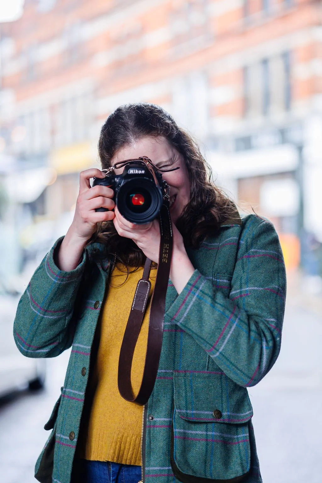 A woman with hair tied back is taking a photo with a Nikon camera outside on a London city street, wearing a green plaid jacket and a mustard yellow sweater. This is a branding photo by Nastia Photography.