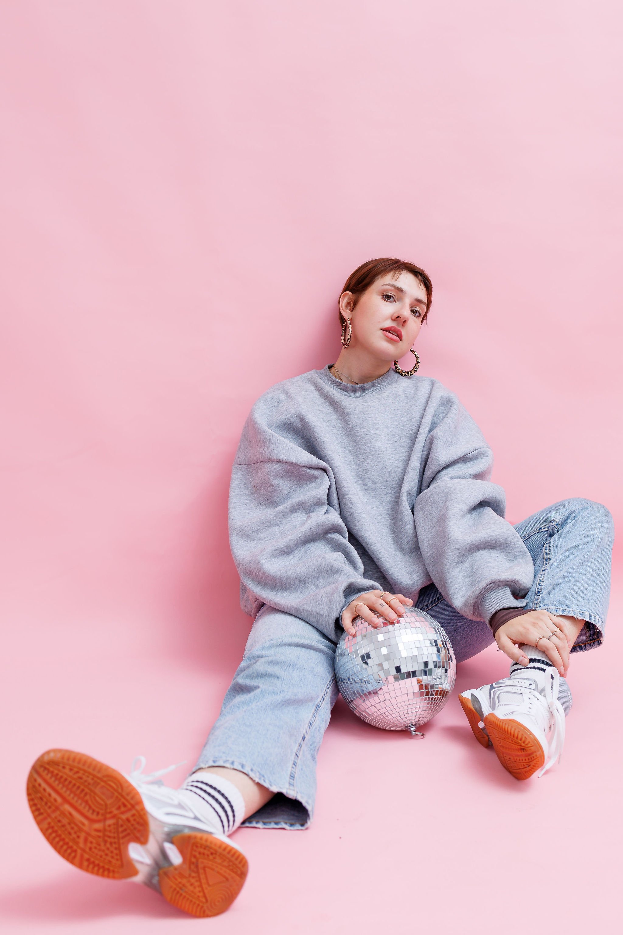 A young woman with short hair in a grey tracksuit, sneakers and hoop earrings, is sitting on the floor against a pink background, holding a disco ball. This is an example of a studio branding photoshoot by Nastia Photography in Kent.