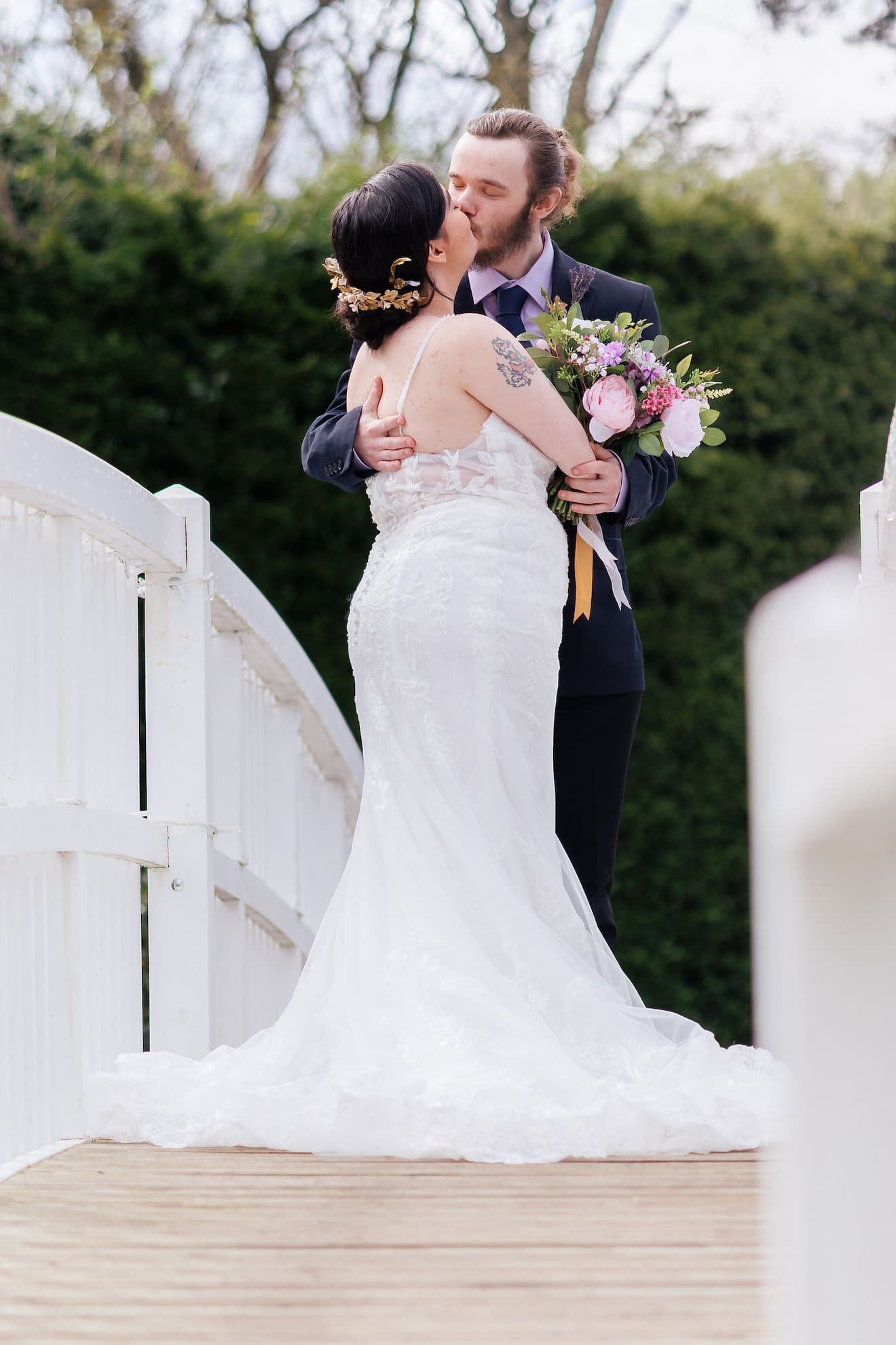 Couple kissing on a bridge, bride in a white wedding gown holding a bouquet, groom in a dark suit, outdoors with trees in the background.
