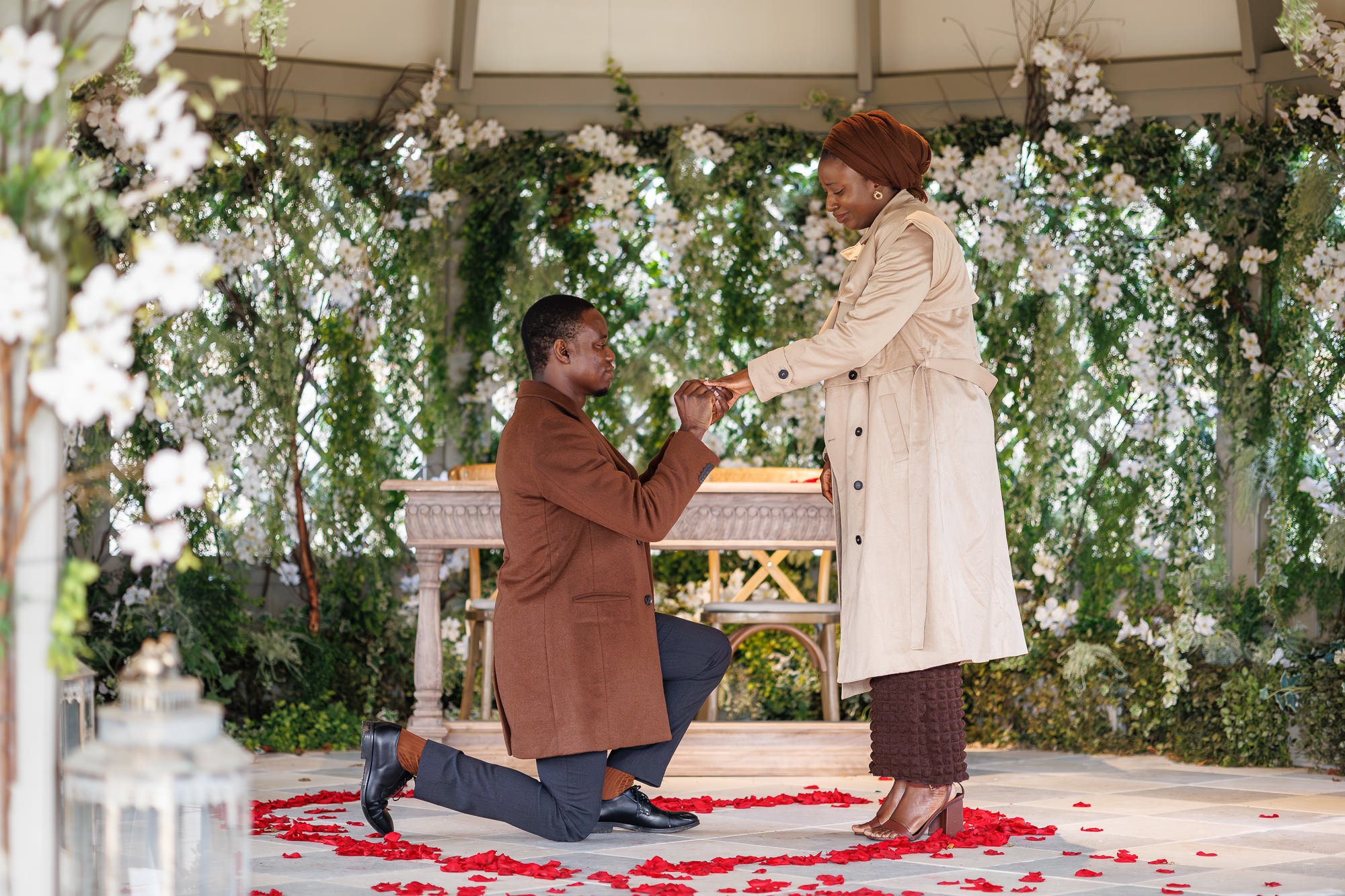 A man proposing marriage to a woman during a wedding ceremony under a decorated arch with white flowers, on a red rose petal-lined floor.
