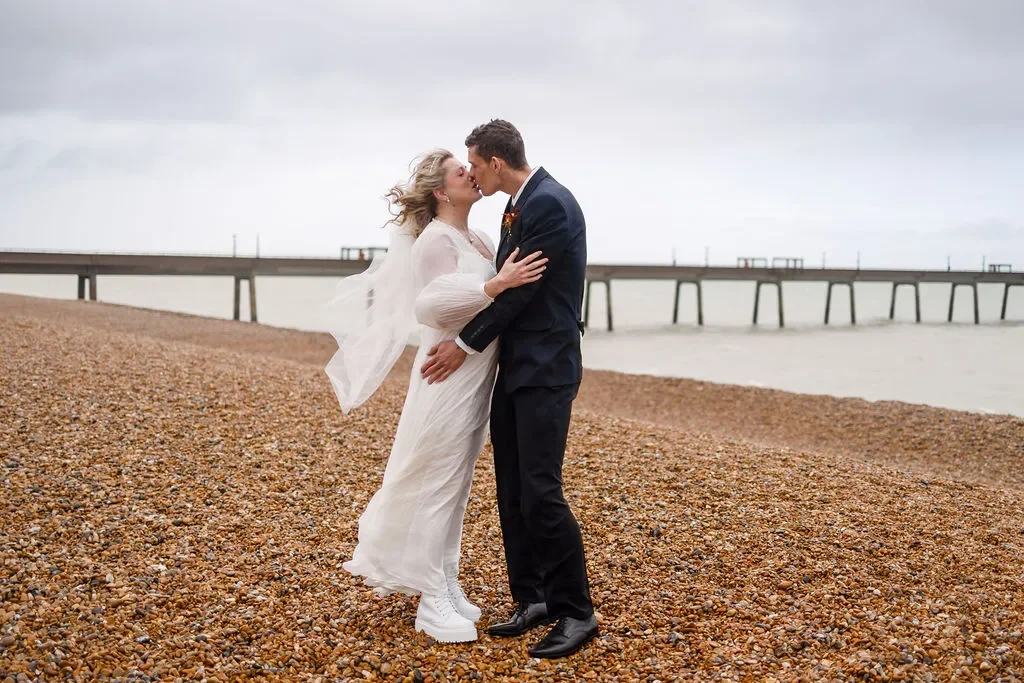 Couple in wedding attire sharing a kiss during a windy day with Deal Pier behind them.