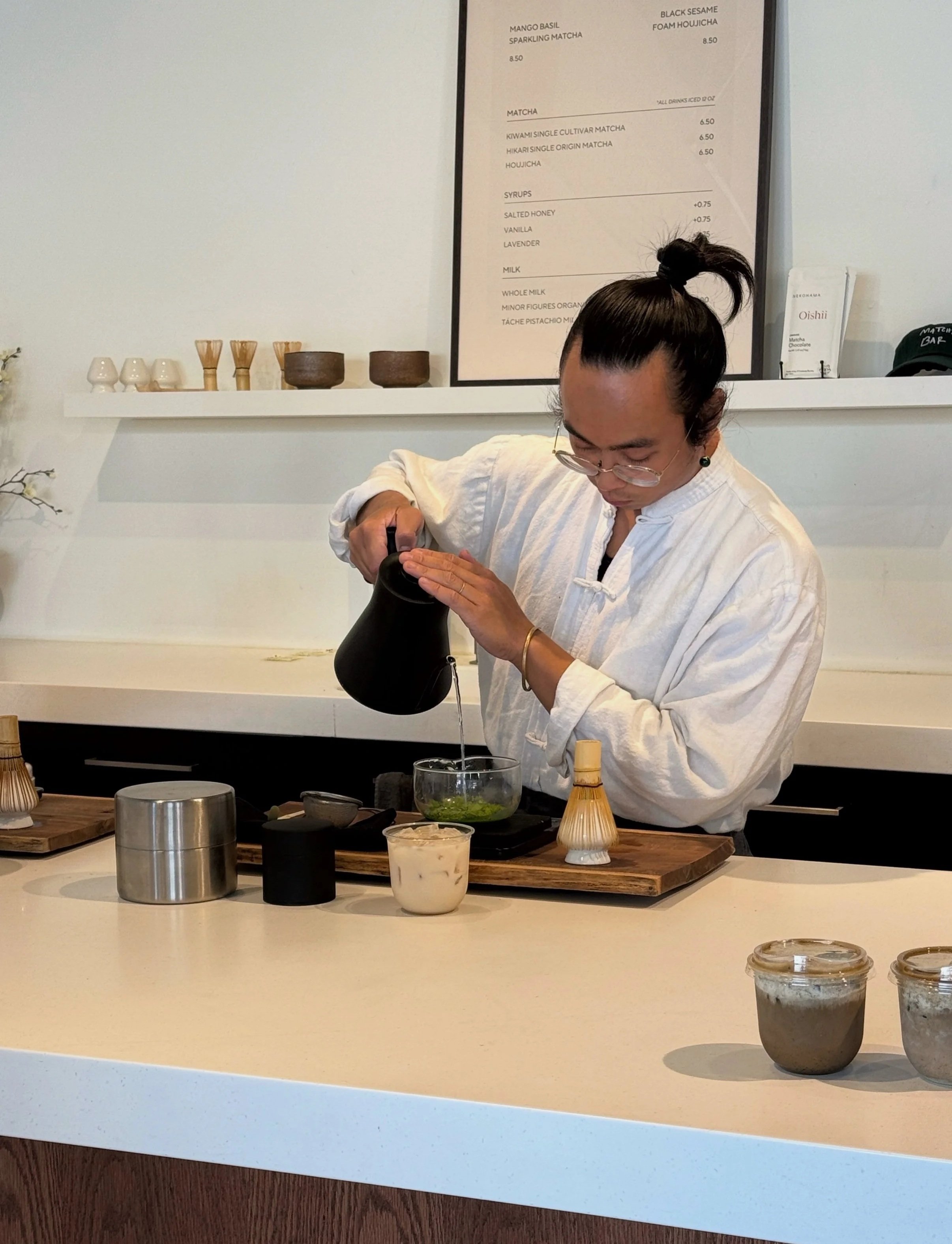 A person with long hair tied up in a bun, wearing glasses and a white shirt, pouring a green liquid from a black kettle into a glass bowl on a counter in a café or tea shop.