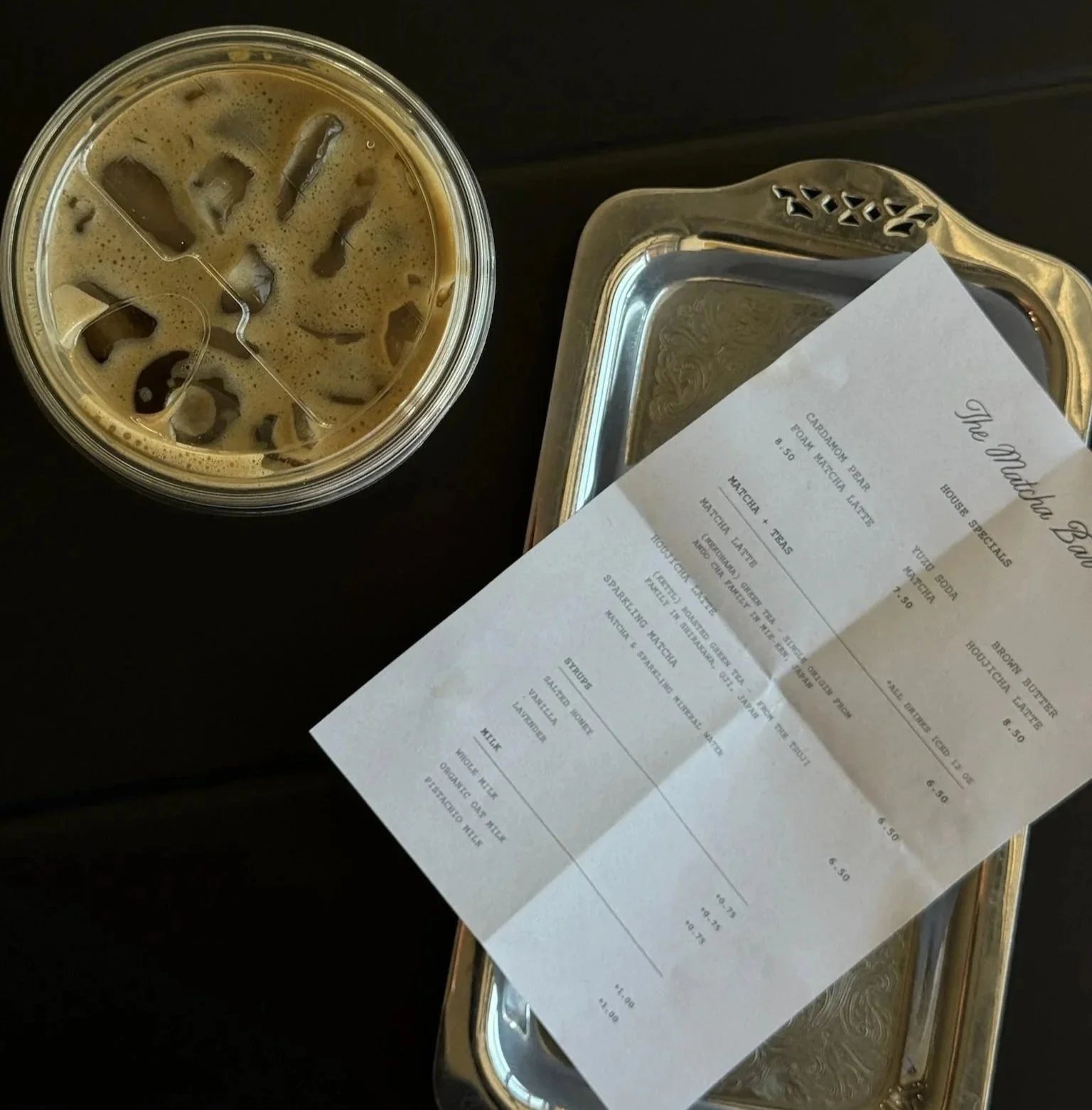 A glass of iced coffee on a black table next to a silver tray with a receipt on top.