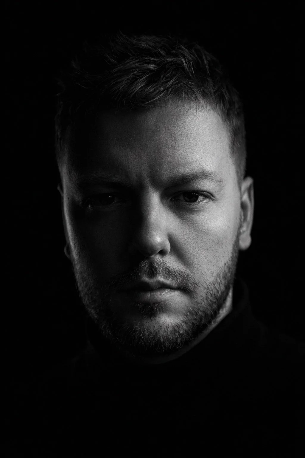 Black and white portrait of a man with short hair and facial hair, looking directly at the camera with a serious expression against a dark background.