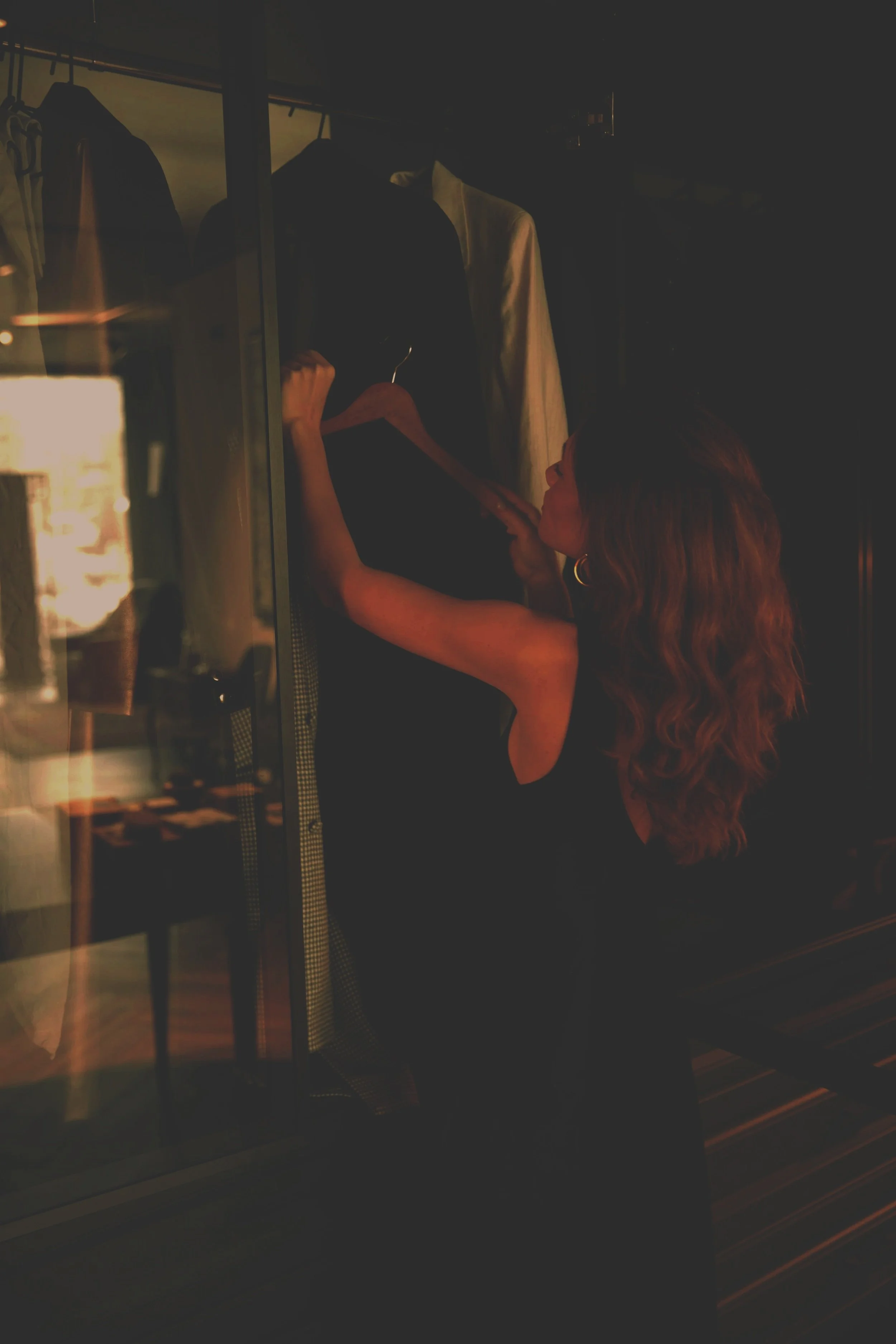 A woman with wavy brown hair, wearing a black dress, is looking at clothing in a dark room, possibly a closet or fitting room, illuminated softly.