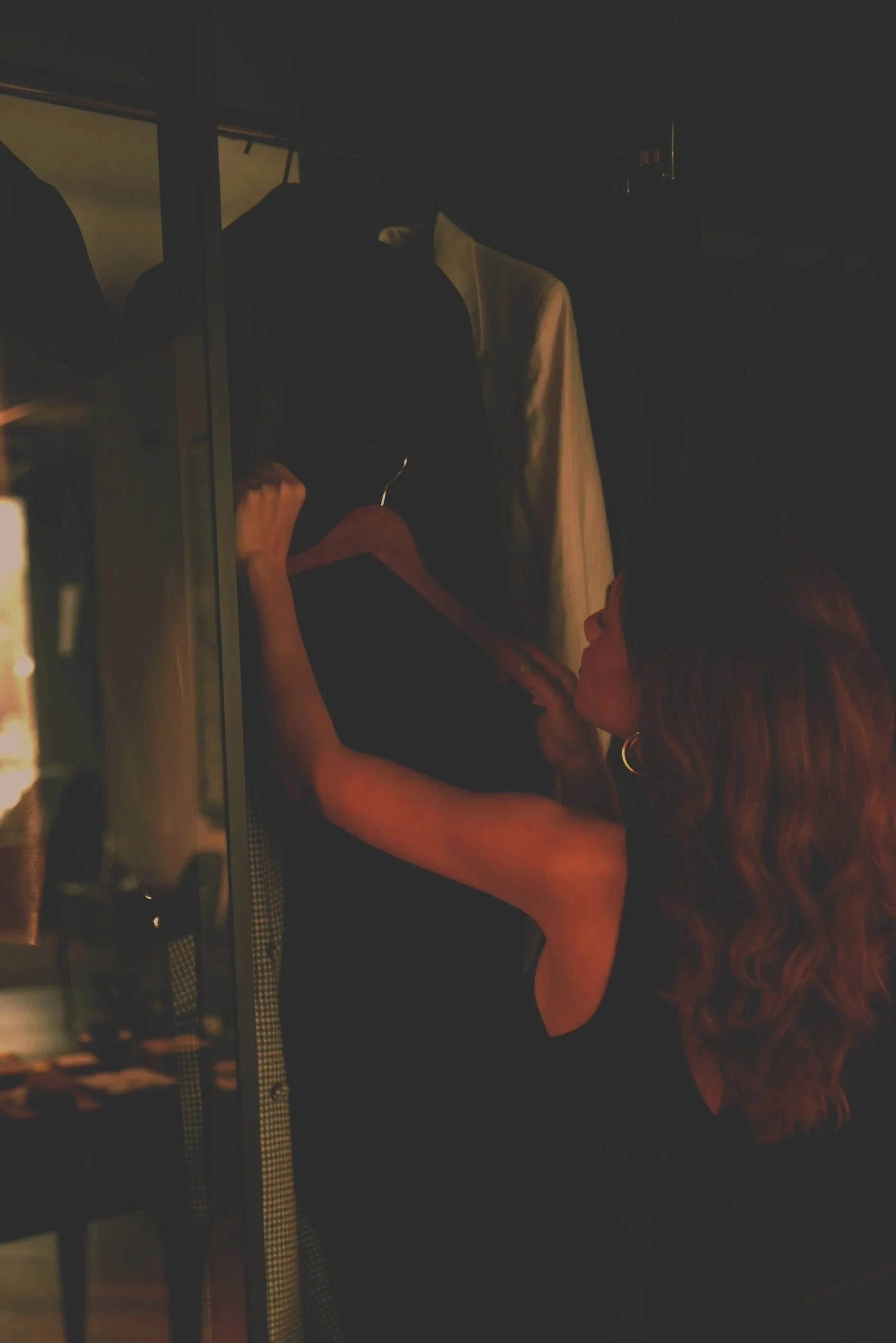 A woman with long wavy hair and hoop earrings is standing in front of a closed closet, holding a black dress on a hanger. She appears to be examining or selecting the dress.