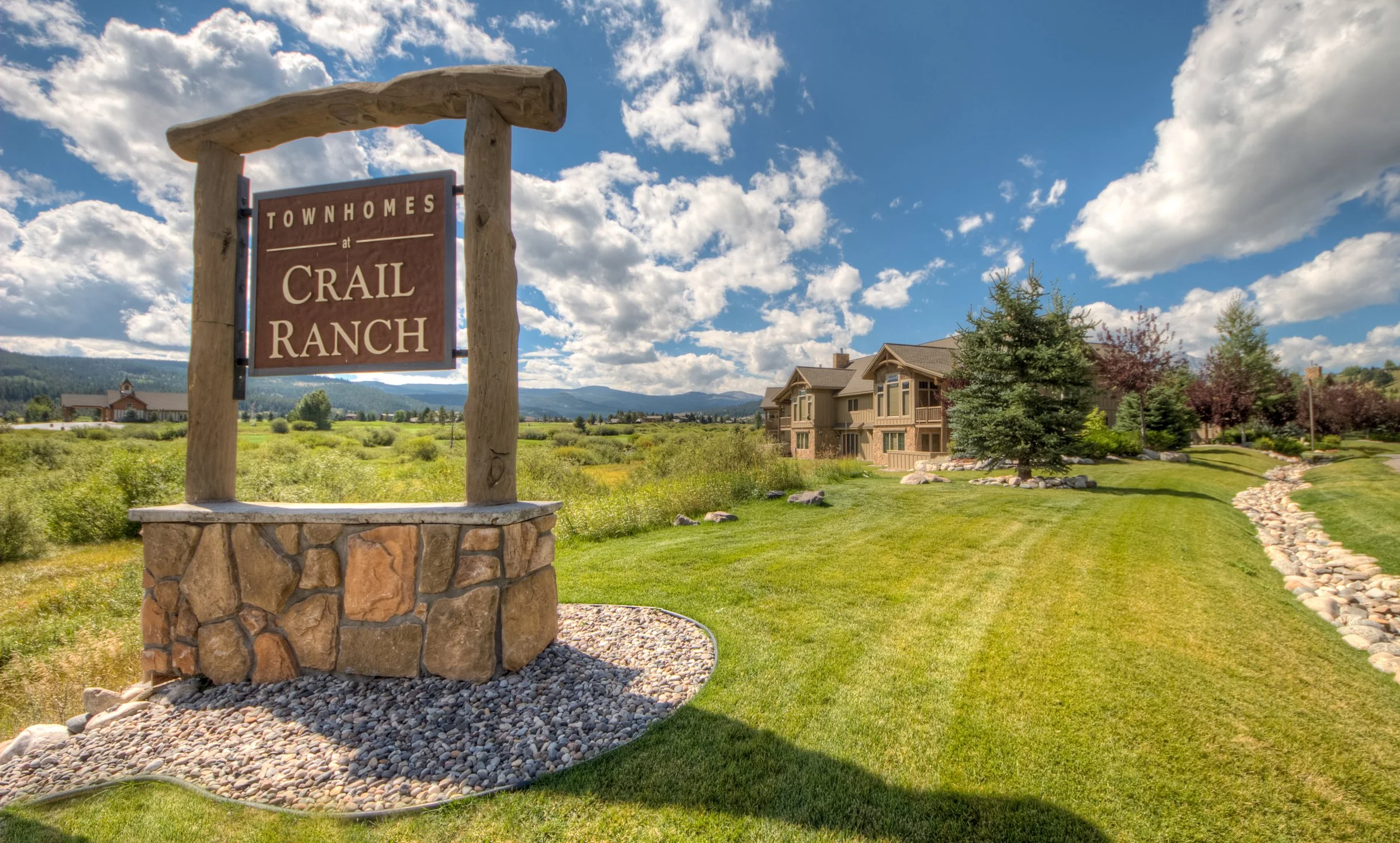 Sign for townhomes at Crail Ranch with a landscaped grass yard, trees, and buildings in the background under a partly cloudy sky.