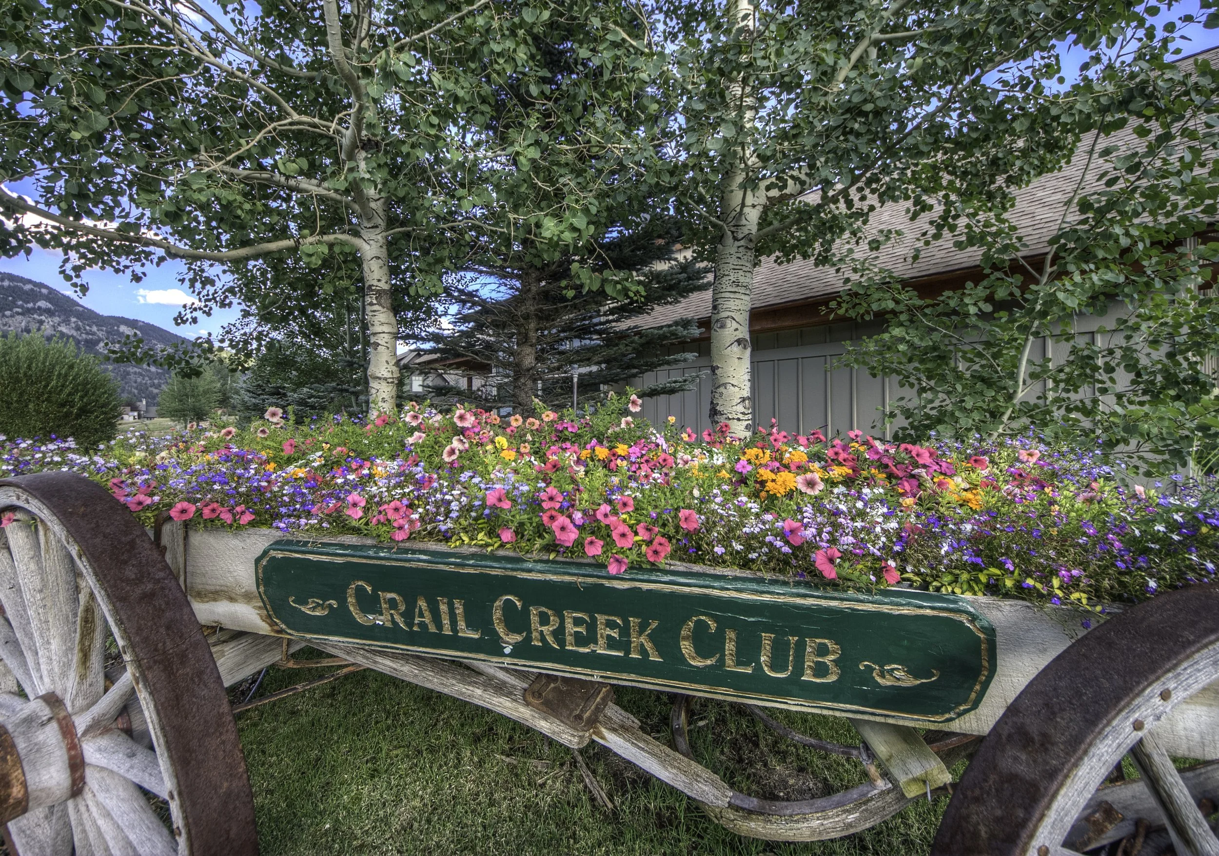 A vintage wooden cart filled with colorful flowers, with a green sign reading 'Crail Creek Club'. Tall trees and mountains are in the background under a blue sky.