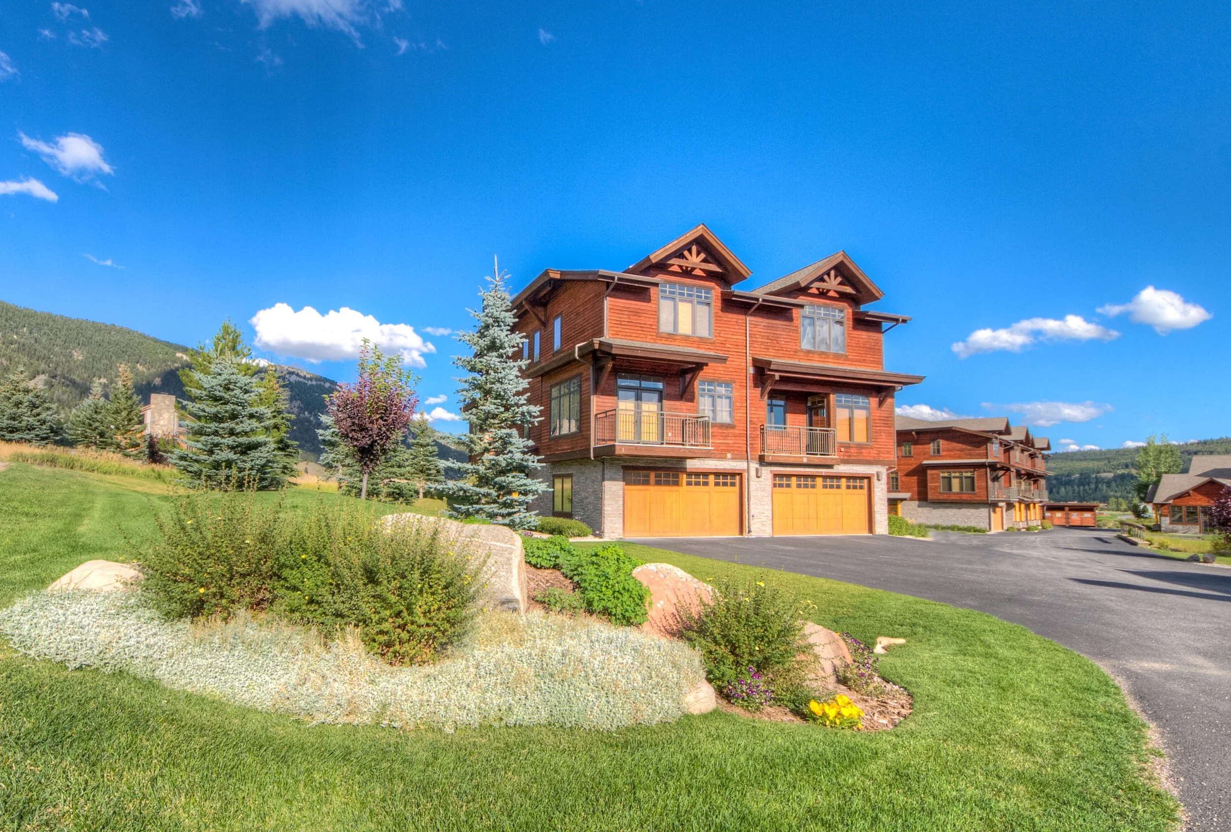 A large, multi-story house made of wood with a landscaped yard, trees, and a paved driveway, under a bright blue sky with scattered white clouds.