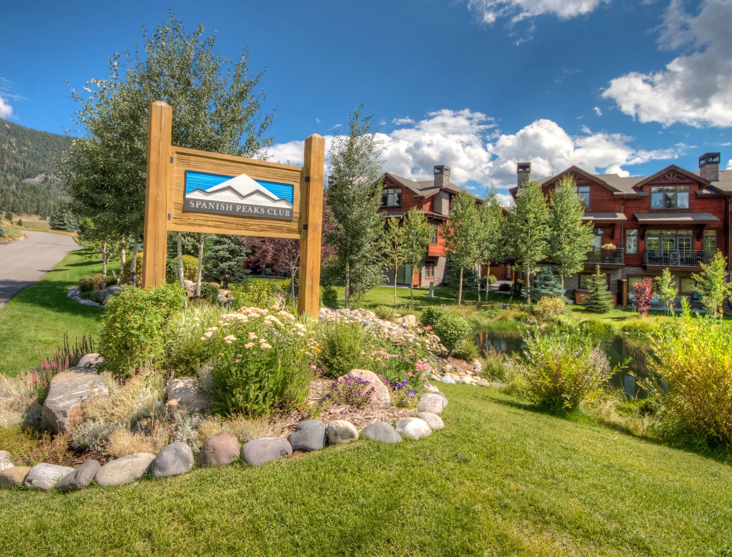 A landscaped area with a sign reading 'Spanish Peaks Club,' surrounded by flowers, rocks, and trees, with a pond and red buildings in the background under a partly cloudy sky.