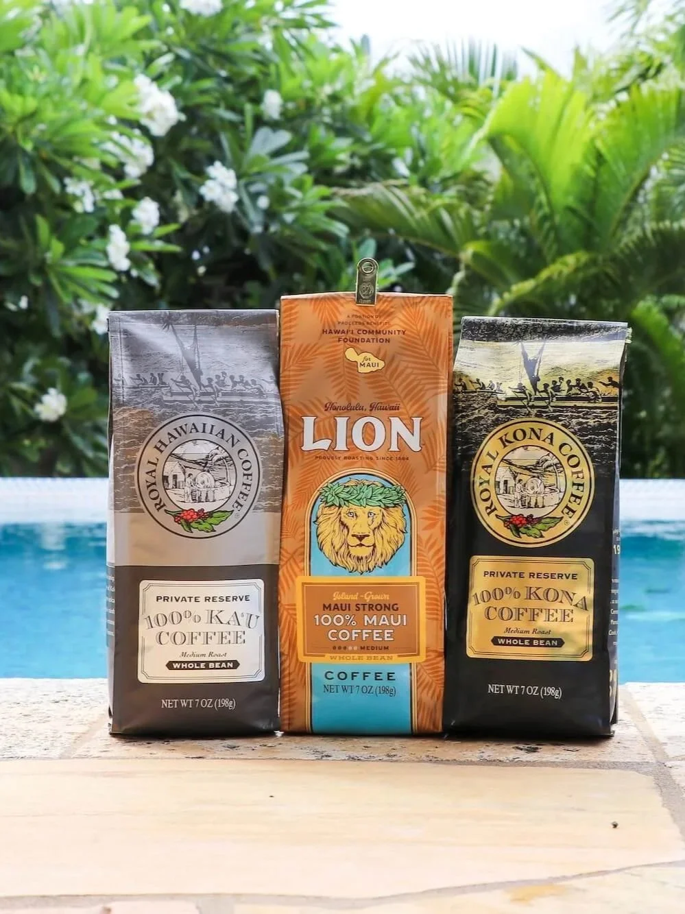 Three bags of Hawaiian coffee sit on a wooden surface with a swimming pool and lush green foliage in the background.