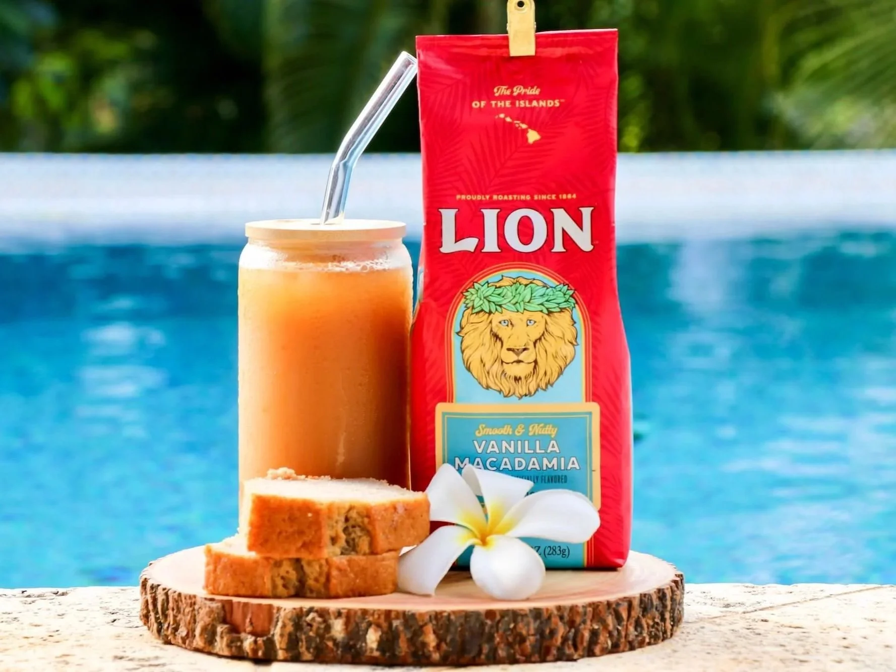 A can of LION vanilla macadamia coffee with a glass of iced coffee and a straw, three slices of blondie dessert, and a white flower on a wooden round tray in front of a swimming pool.