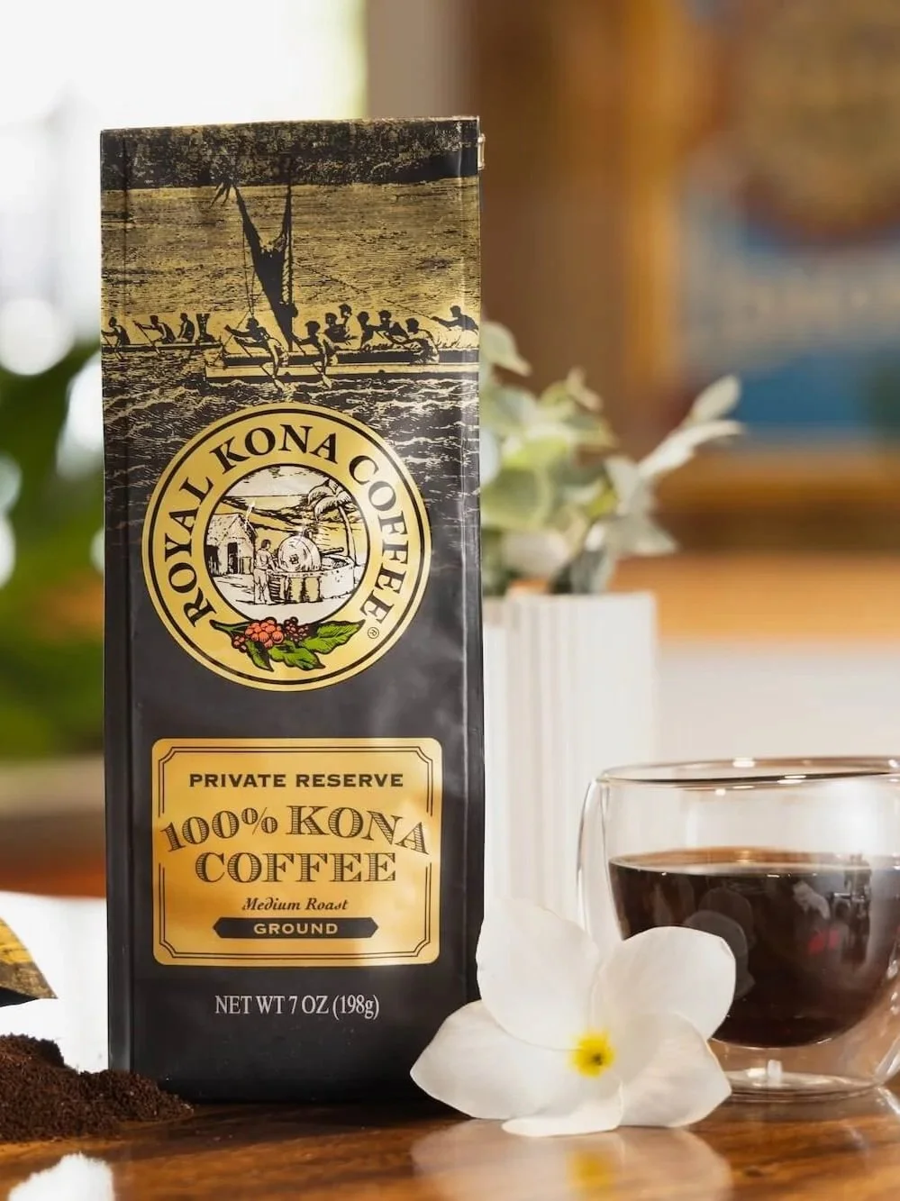 A package of Royal Kona Coffee with a glass of brewed coffee and a white flower next to it on a wooden surface.