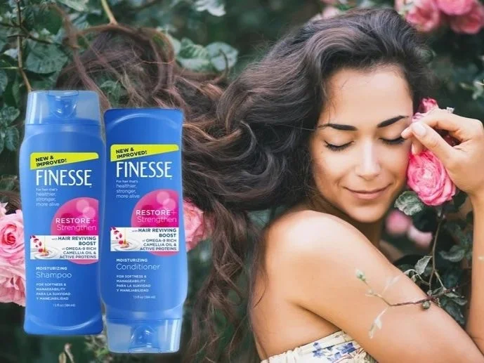 A woman with long, curly hair holding a pink flower, outdoors with greenery and pink flowers in the background, with two Finesse hair care product bottles in the foreground.