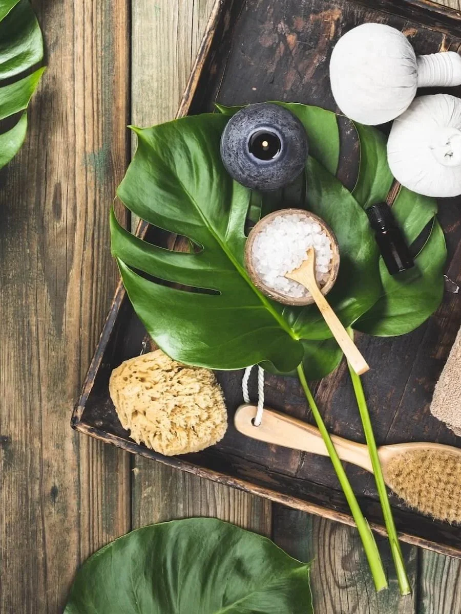 Spa or skincare setting with green tropical leaves, massage stones, a wooden scrub brush, a small bowl of salts with a spoon, a black essential oil bottle, and two white massage balls on a rustic wooden tray.
