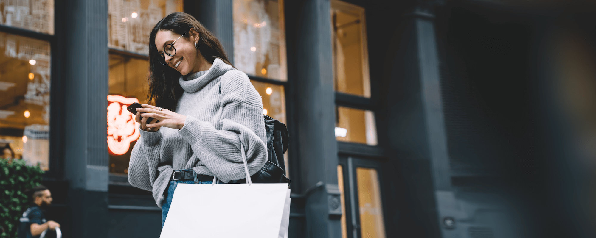 A woman with glasses, wearing a gray turtleneck sweater, looking at her smartphone and smiling, outside a building with large windows and a neon sign in the background.