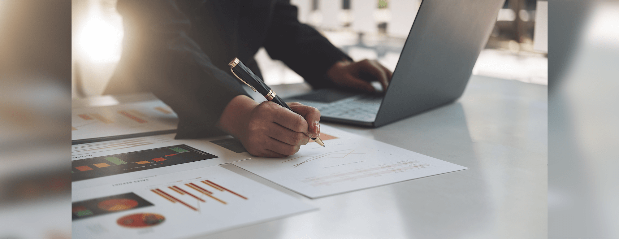 Person working at a desk with a laptop, analyzing printed charts and graphs, holding a pen for note-taking.