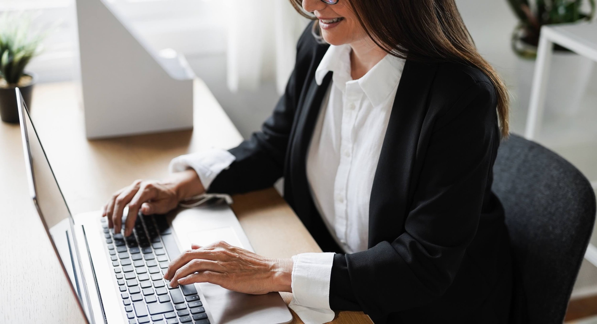A woman in a black suit and white shirt working on a laptop at a desk in an office.