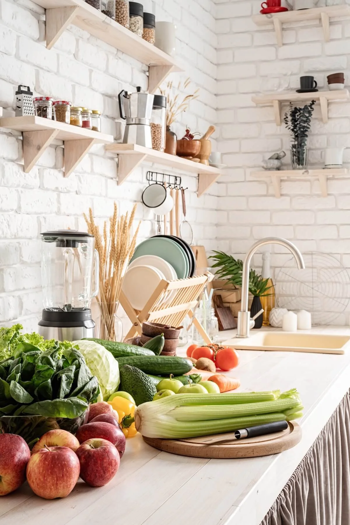 Fresh vegetables like apples, lettuce, cucumbers, tomatoes, celery, and carrots arranged on a kitchen counter with a white brick wall background, open shelves, a blender, and kitchen utensils.