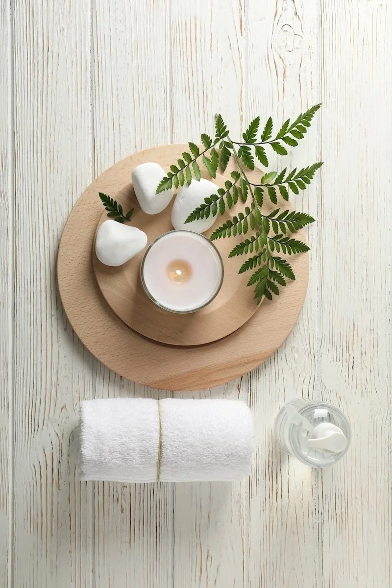 Spa or relaxation setup with a lit candle, white stones, fern leaves on a round wooden tray, a set of rolled white towels, and a spray bottle on a light wooden surface.