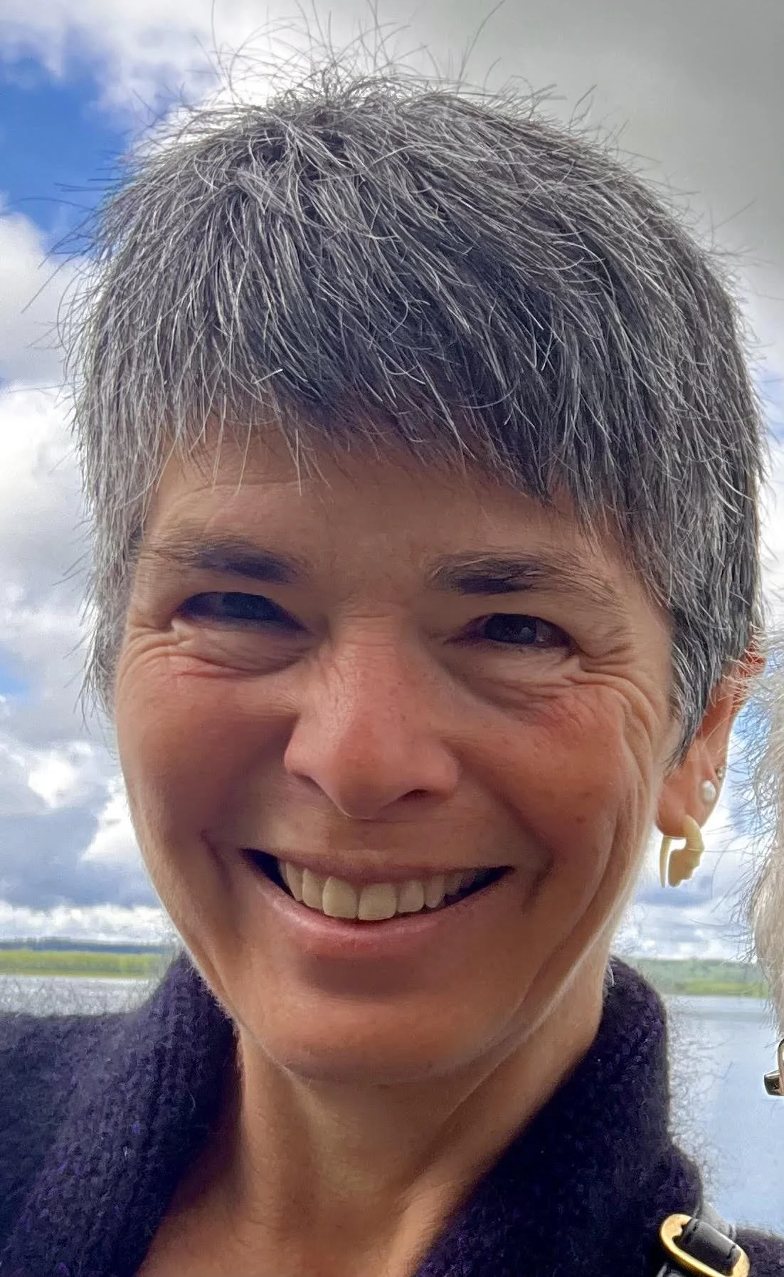 Close-up of a smiling woman with short gray hair and a dimple, outdoors with cloudy sky and water in the background.