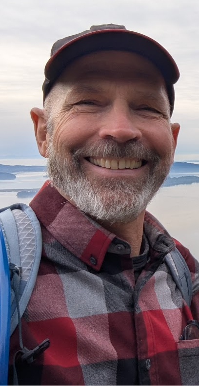 A smiling man with a beard and mustache wearing a cap and plaid shirt, outdoors with a water body and cloudy sky in the background.