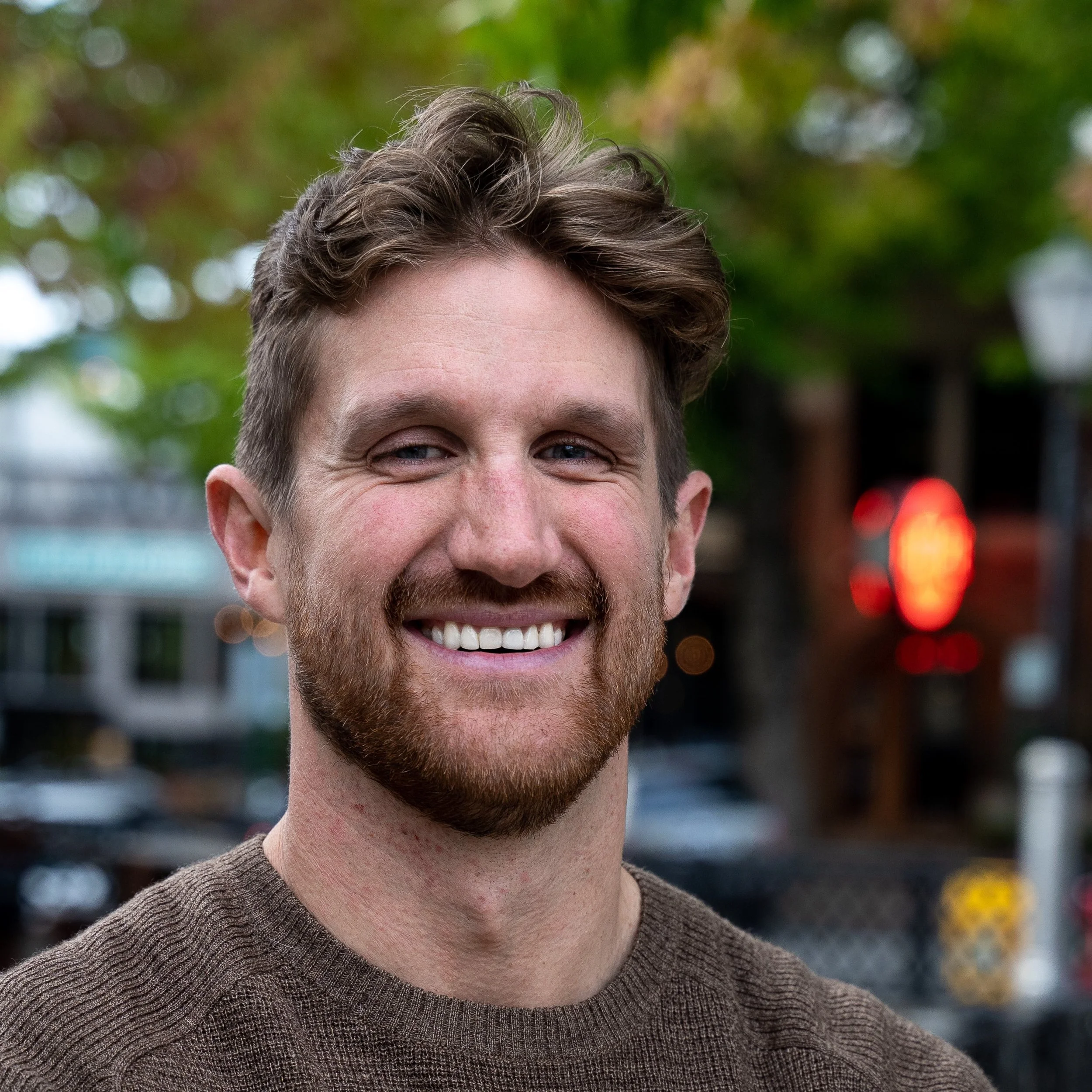 A smiling man with a beard and short wavy hair outdoors with trees and blurred lights in the background.