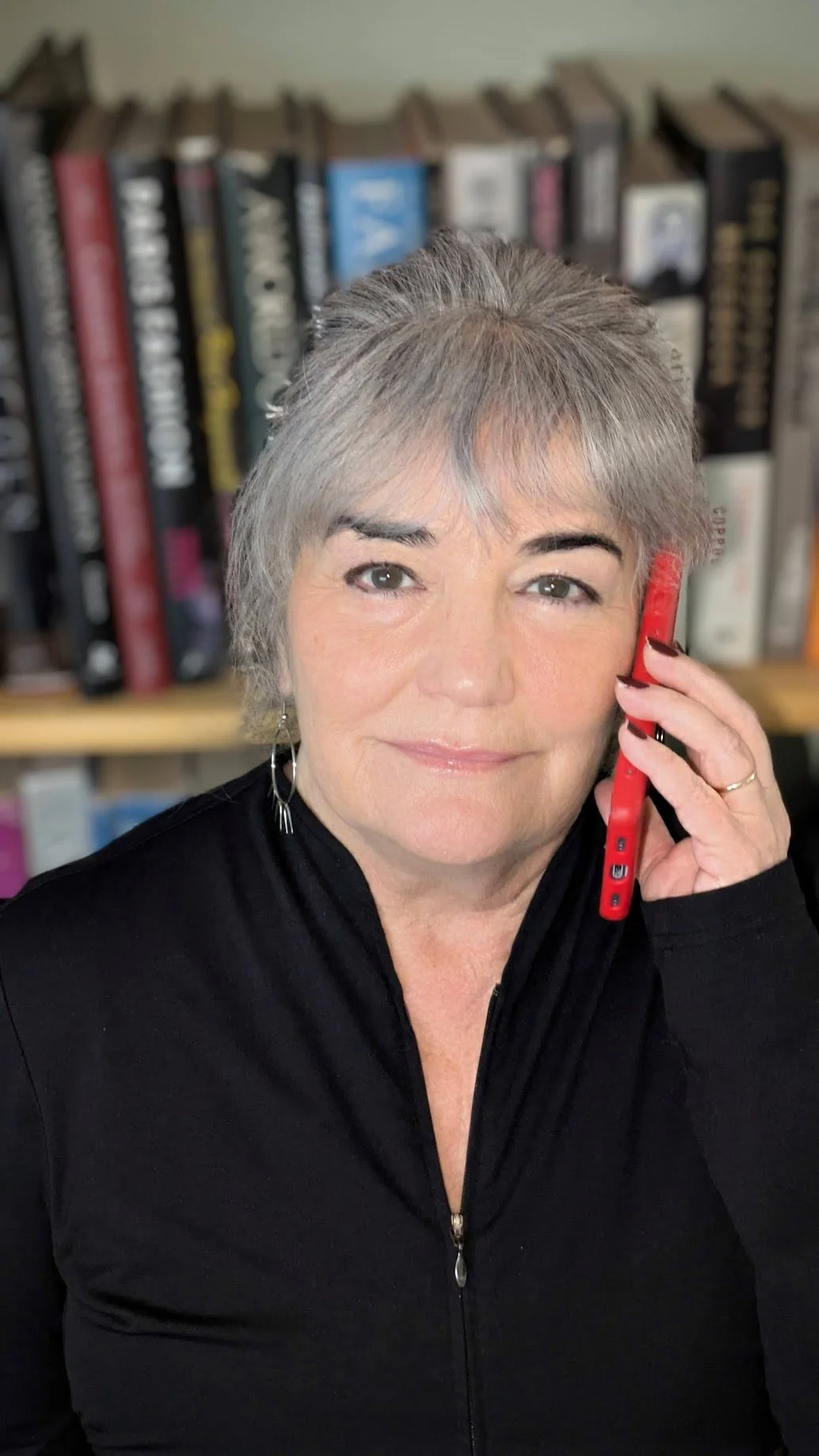 A middle-aged woman with short gray hair talking on a red cellphone, wearing a black zip-up top, with a bookshelf in the background.
