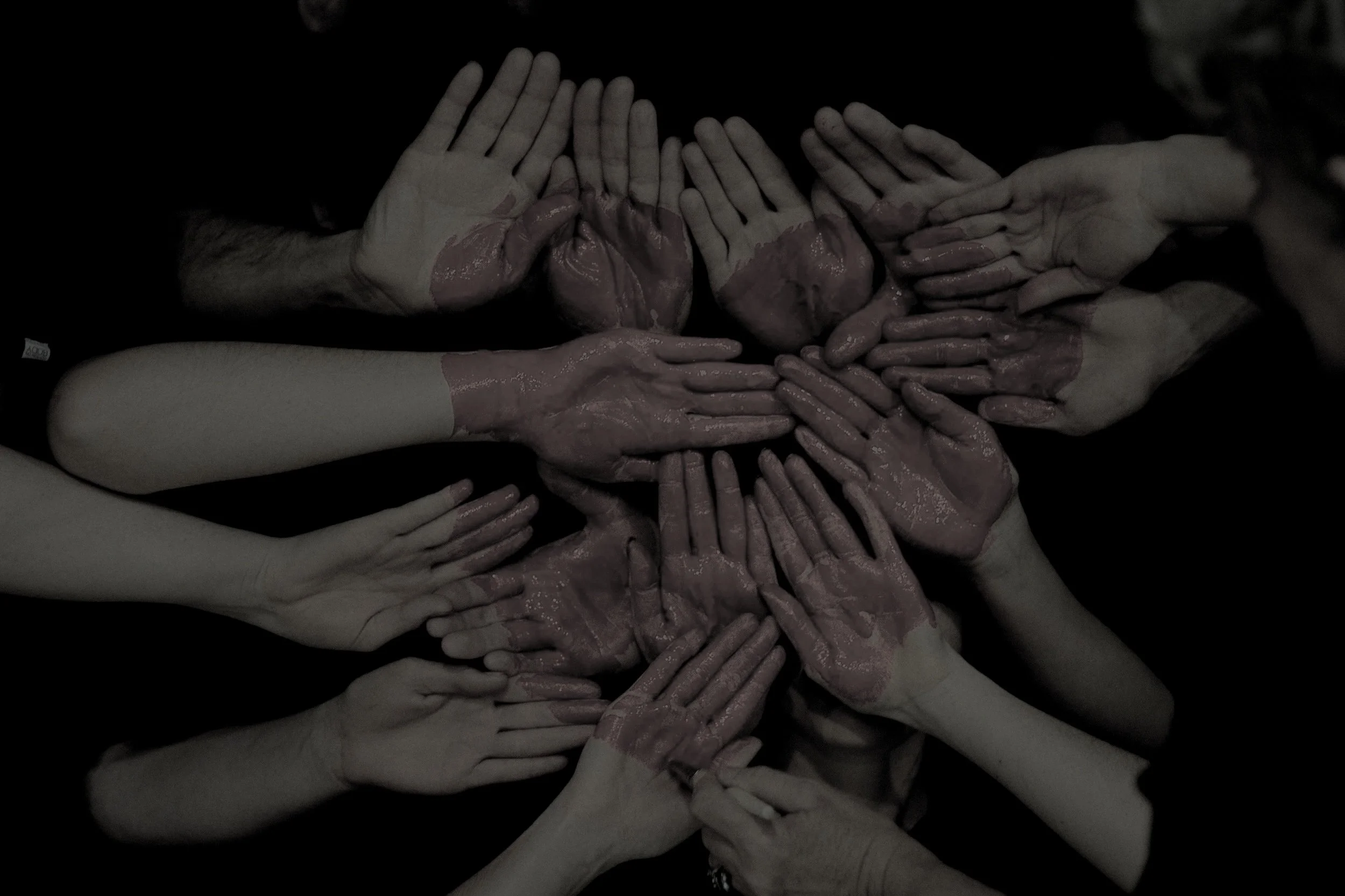 Multiple hands covered in pink paint arranged to form a heart shape against a black background.