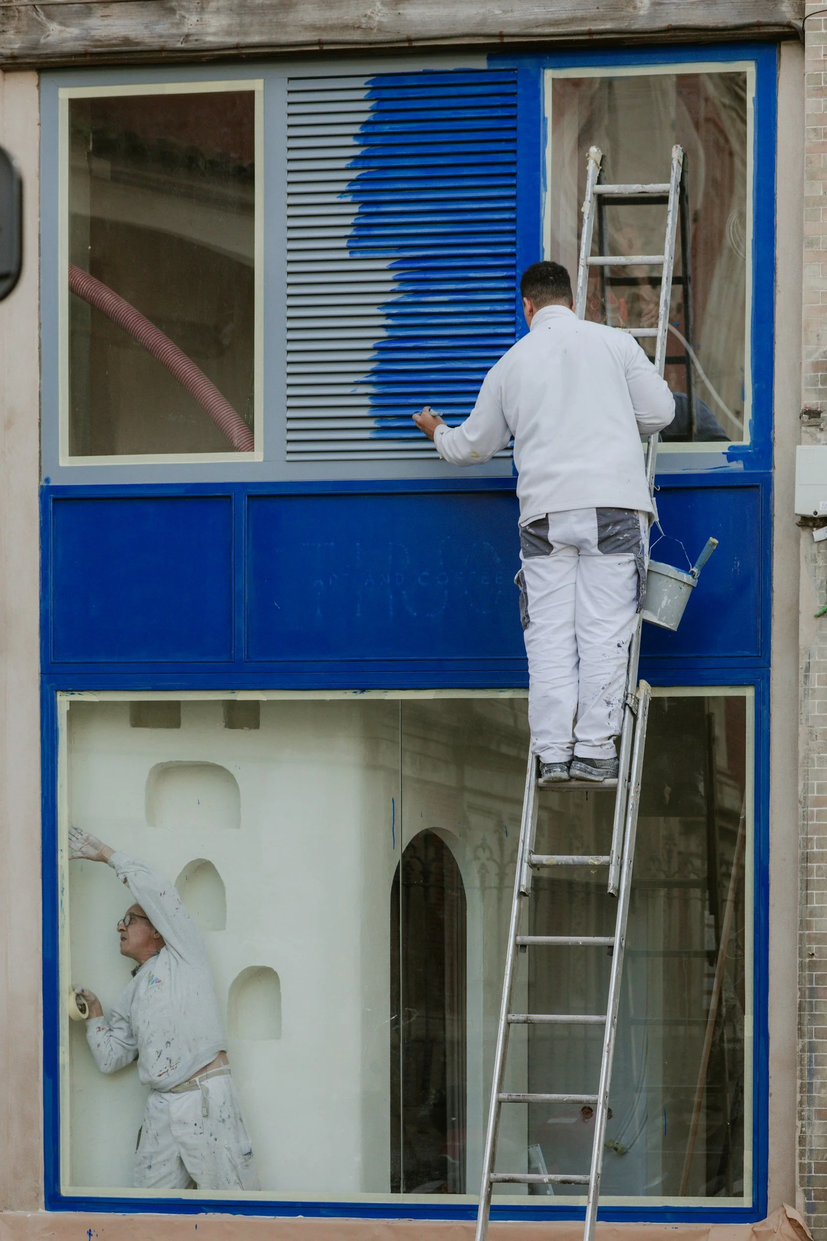 Two workers painting a building. An man on a ladder painting blue horizontal panels on the upper portion, and a woman on the ground painting white on the lower portion.