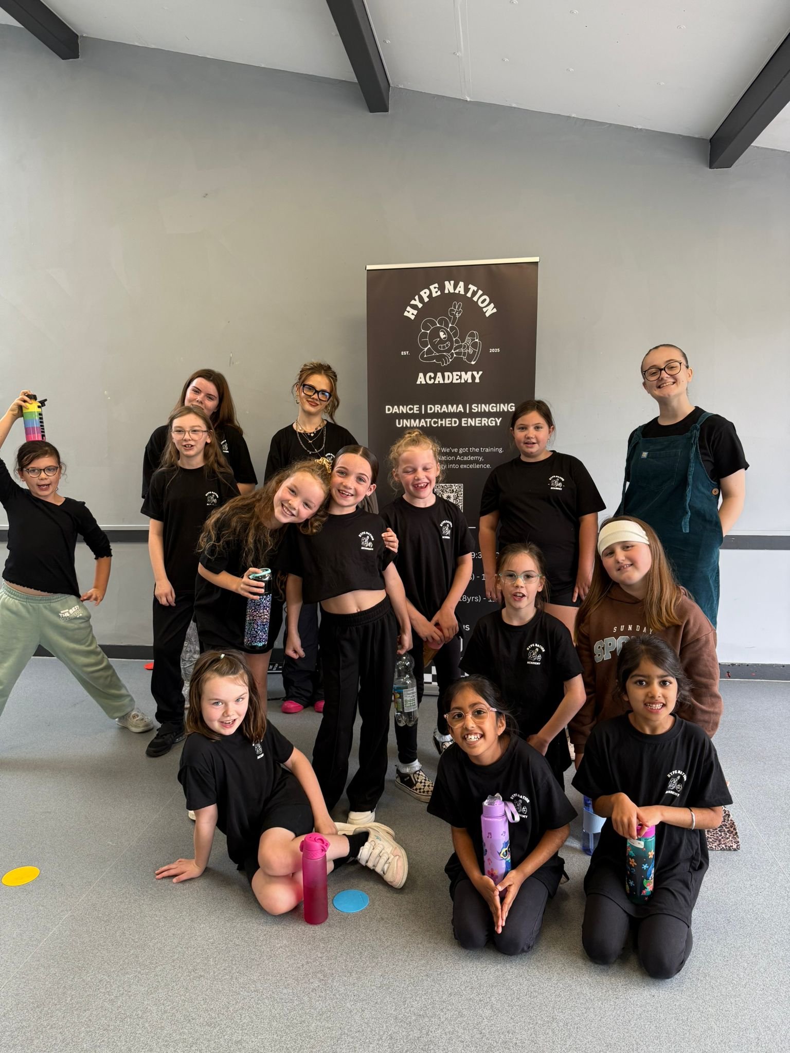 Group of young girls and two adults in black t-shirts with a 'Hype Nation Academy' banner in the background, posing for a photo indoors.