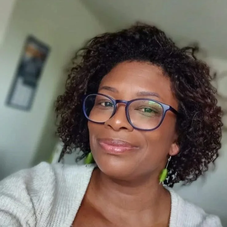 A woman with curly hair wearing glasses and earrings, smiling at the camera.