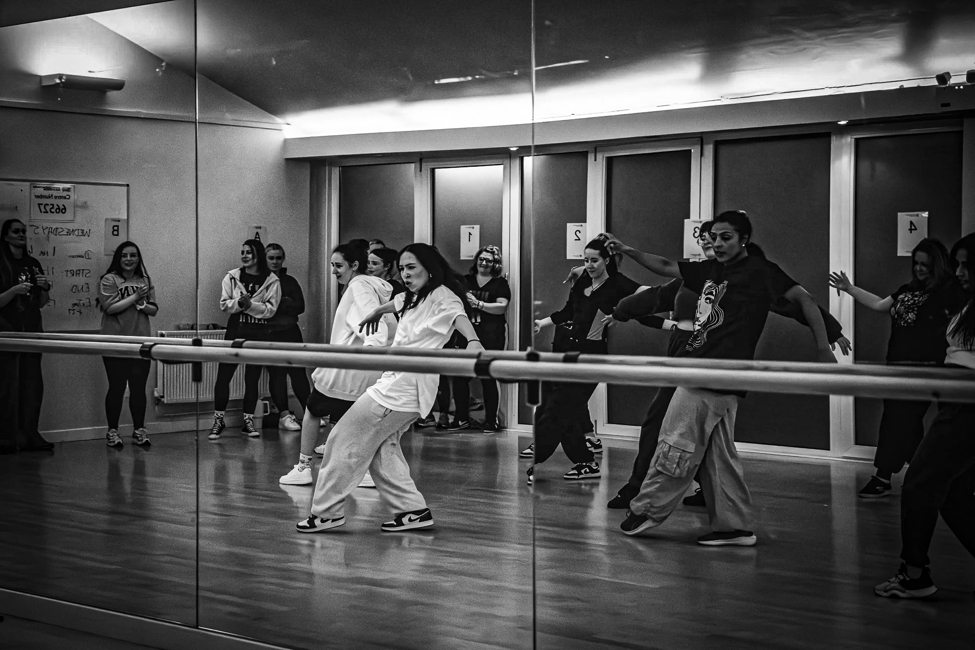 A group of people dancing in a dance studio with mirrors on the wall, some wearing casual clothes and sneakers, with a ballet barre in the foreground.