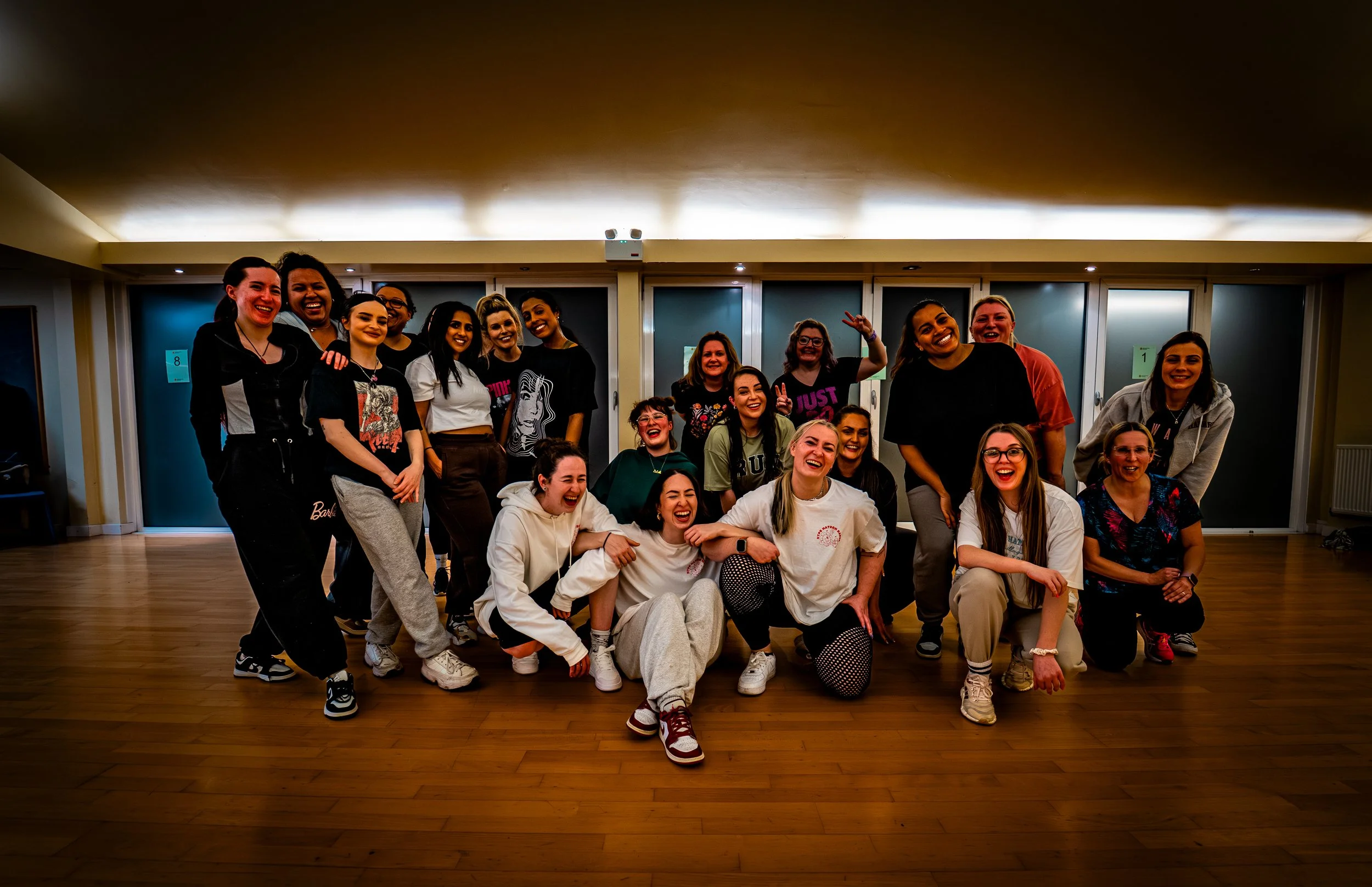 Group of smiling women posing together indoors, some kneeling and others standing, with wooden floor and large windows in the background.