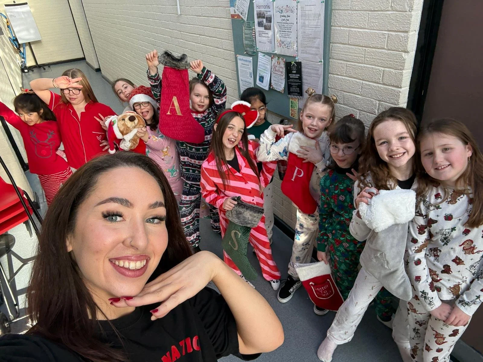 A group of children and a woman taking a selfie together in Christmas pajamas, with some children holding Christmas stockings and a small stuffed dog, in a hallway decorated for the holidays.