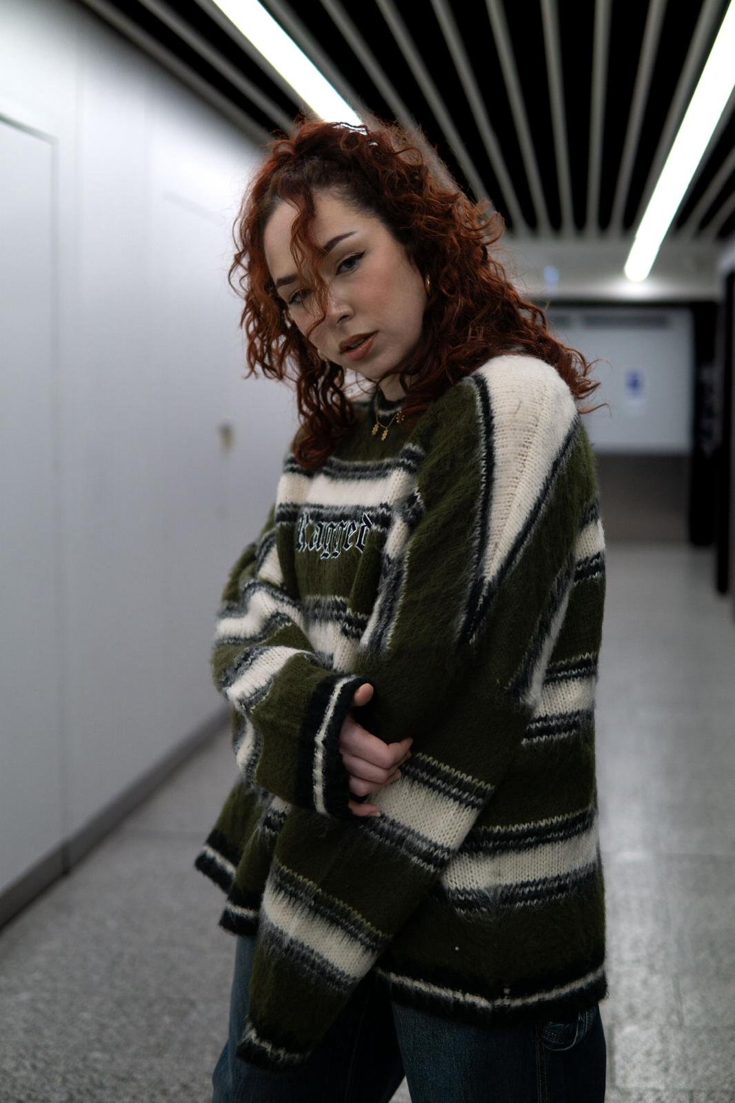 A young woman with curly red hair, wearing a green and white striped sweater, stands in an indoor hallway with a white wall and ceiling with black and white striped panels.