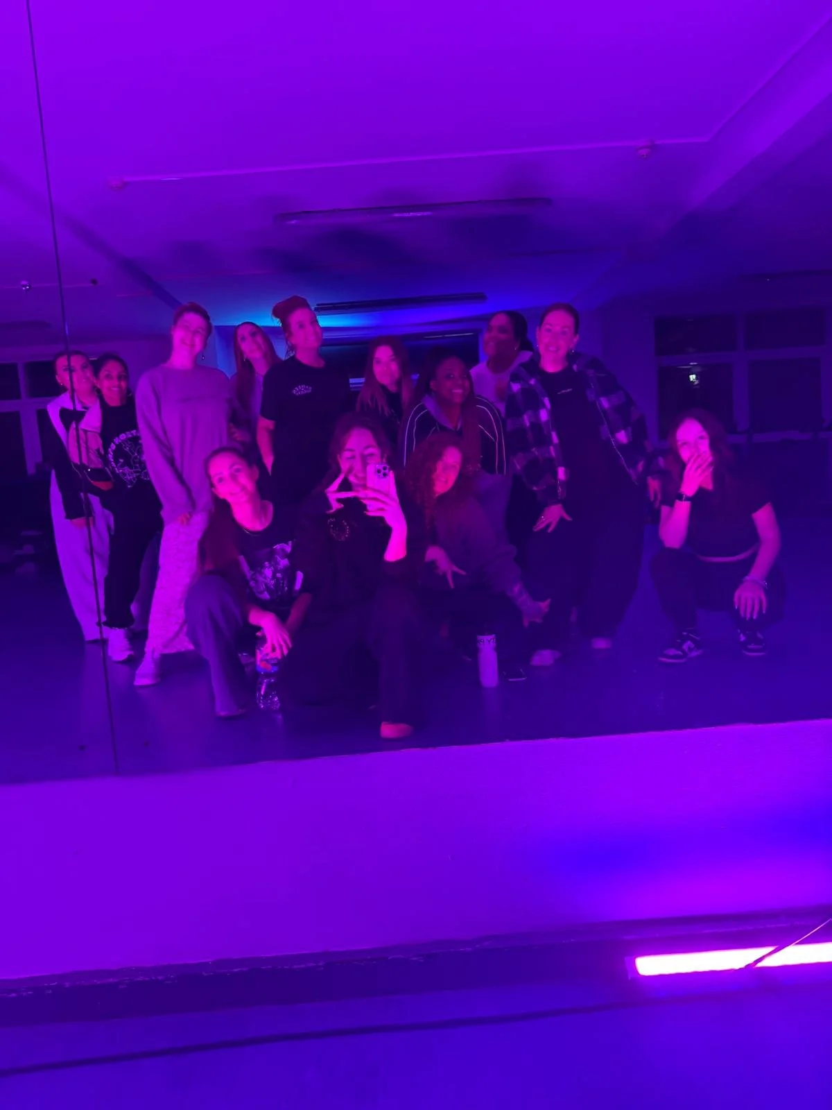 A group of people taking a selfie in a room illuminated with purple and blue lights, some standing and some squatting, with a reflective wall behind them.