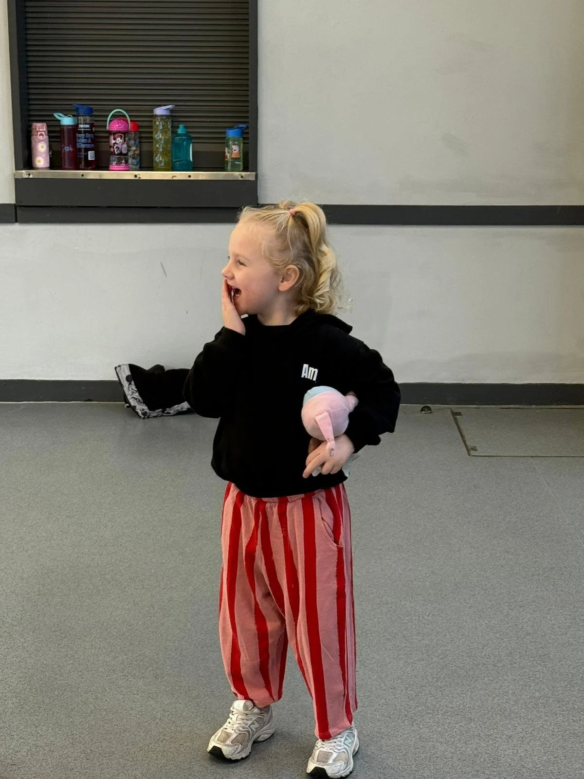 A young girl with blonde curly hair in a high ponytail is standing on a gray carpeted floor, laughing and covering her mouth with her right hand. She is wearing a black hoodie, red and pink striped pants, and white sneakers. She is holding a pink and blue stuffed toy in her left hand. The background features a gray wall with a small window or vent with bottles on a ledge beneath it.