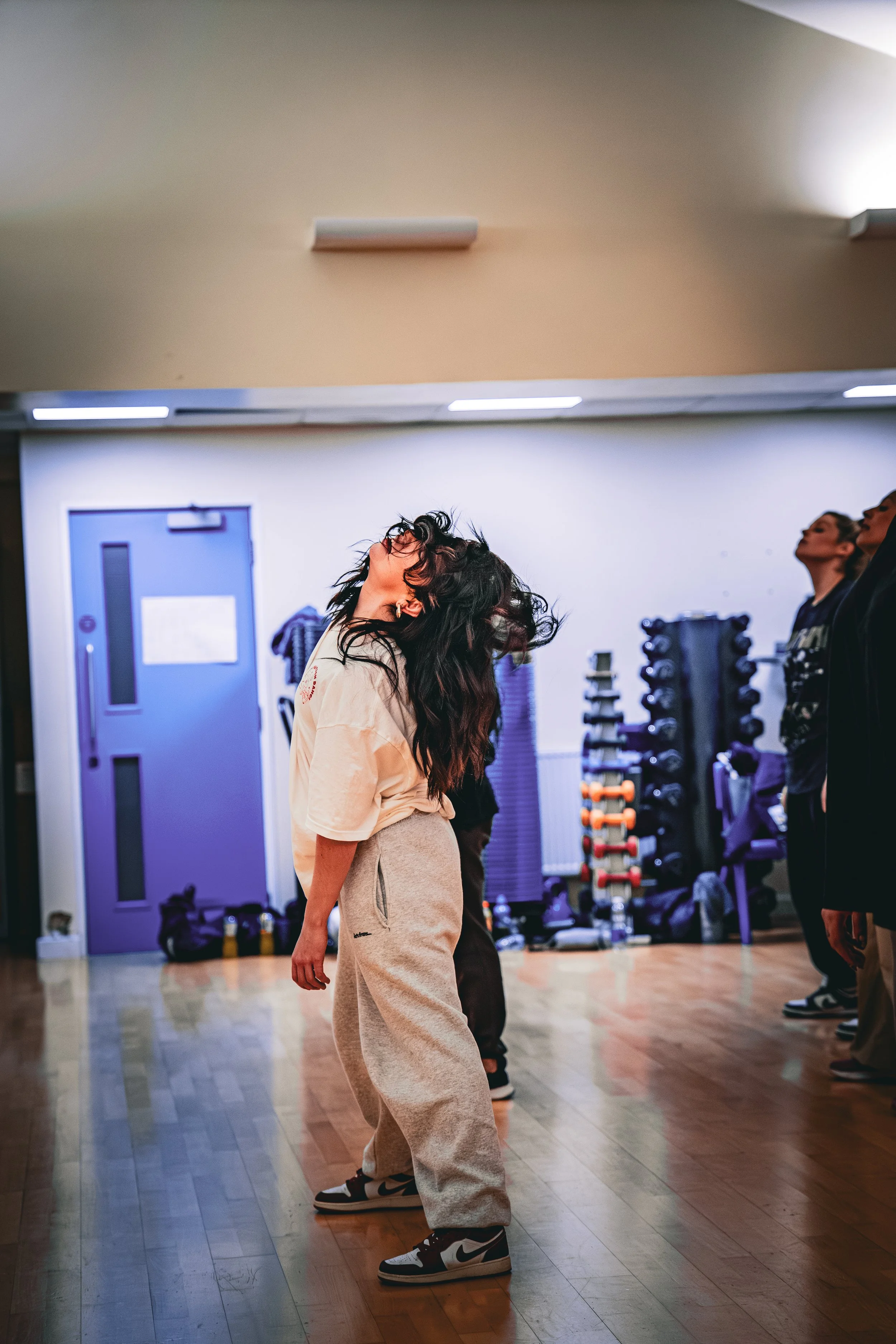 A woman with long dark hair dancing intensely with her head tilted back in a dance studio, with people in the background and exercise equipment against the wall.
