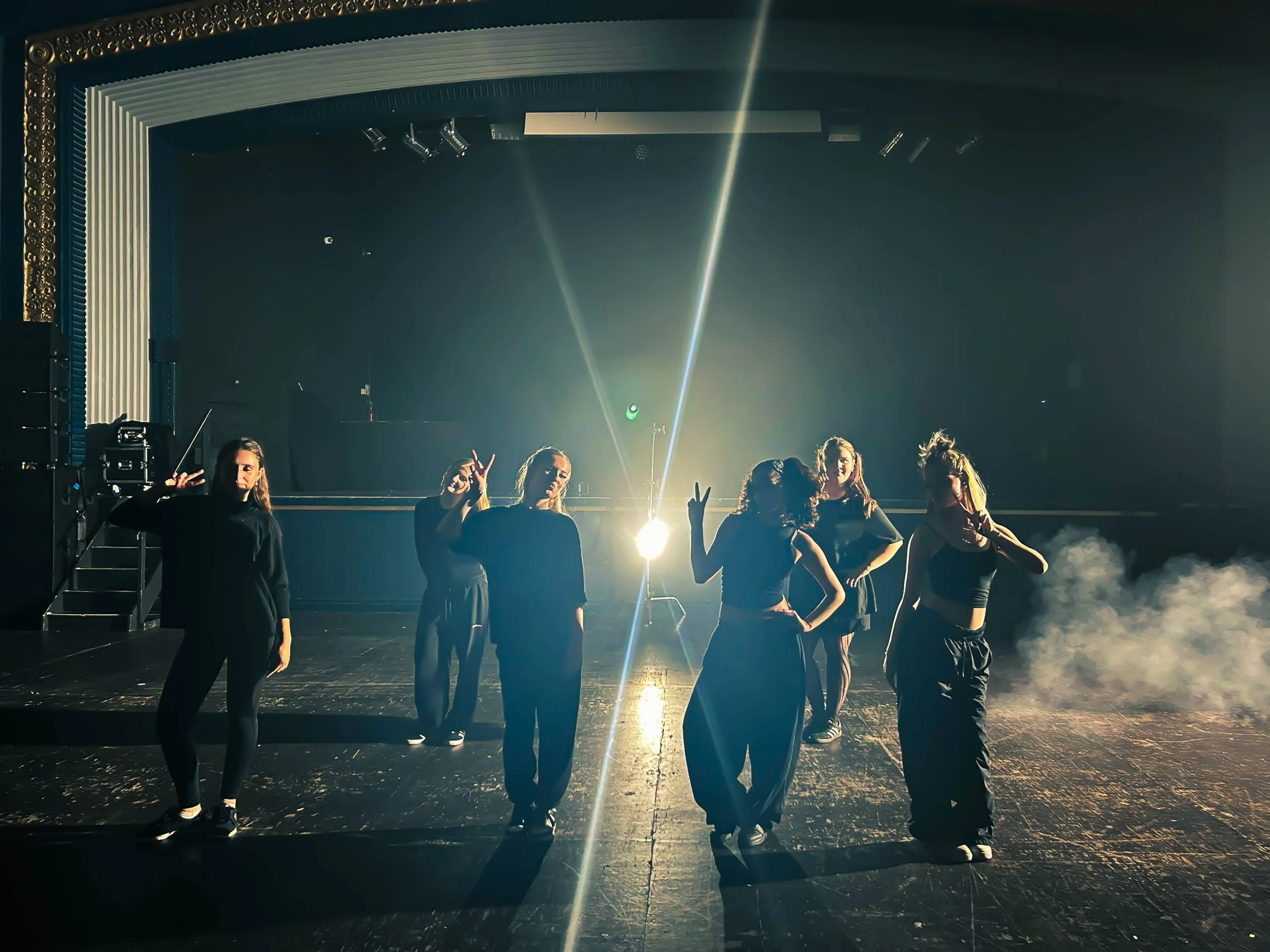 Six women in black outfits posing on a stage with bright backlight and smoke effects.