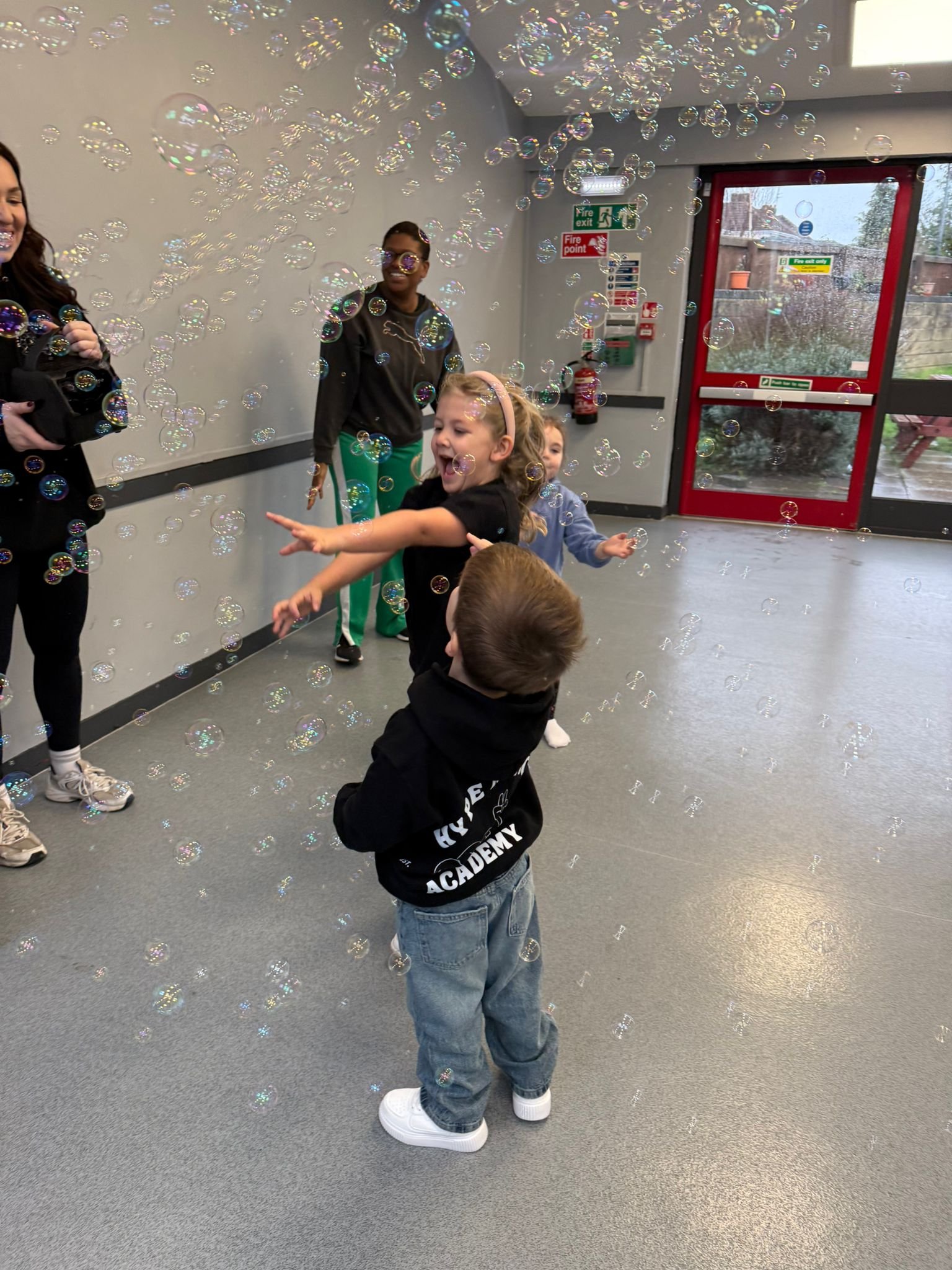 Kids playing with soap bubbles indoors, with two adults holding bubble wands, and a red exit door visible in the background.