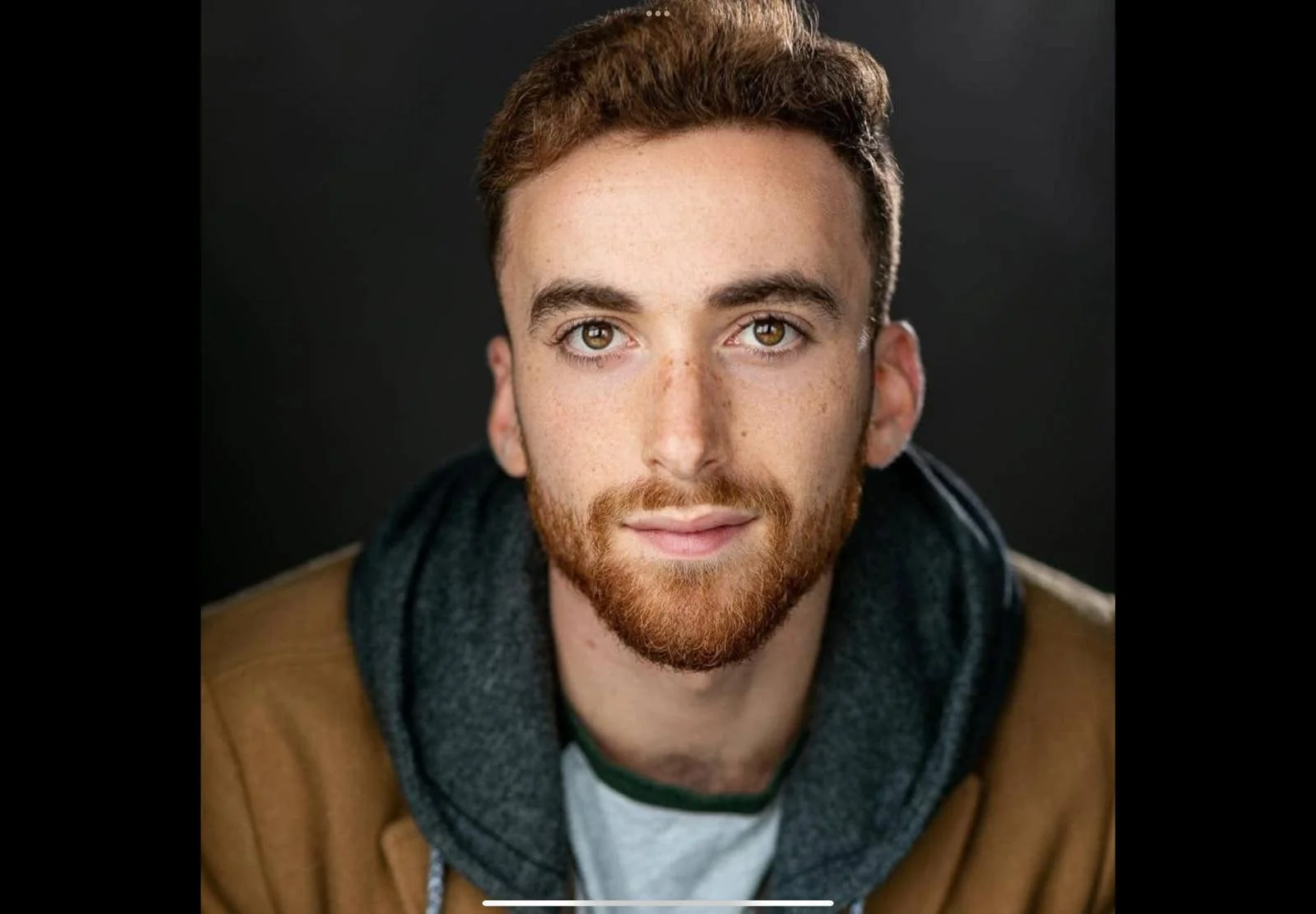 Close-up portrait of a young man with short brown hair, green eyes, and a red beard, wearing a brown jacket and a grey hoodie, against a black background.
